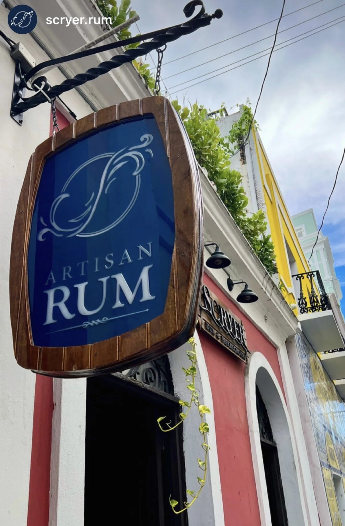 Outdoor signboard for Artisan Rum with a blue background and white text, mounted on a building with red, white, and yellow walls, graffiti-style black metal accents, and a plant hanging from the roof.
