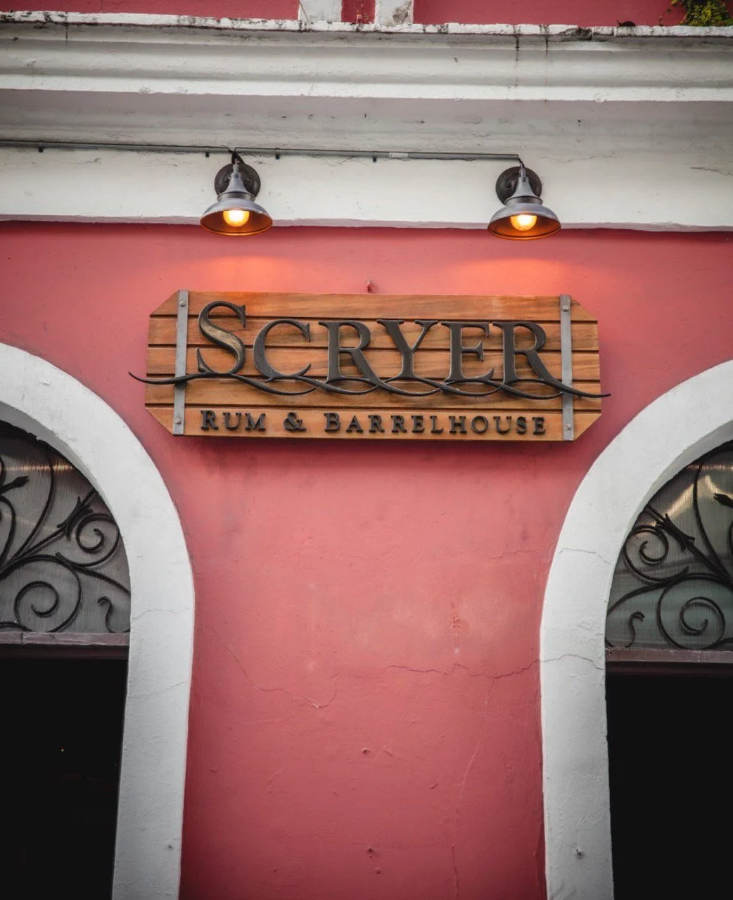 A wooden sign reading 'ISCRYER Rum & Barrelhouse' mounted on a pink wall above an arched window with black wrought iron bars, illuminated by two small lamps.