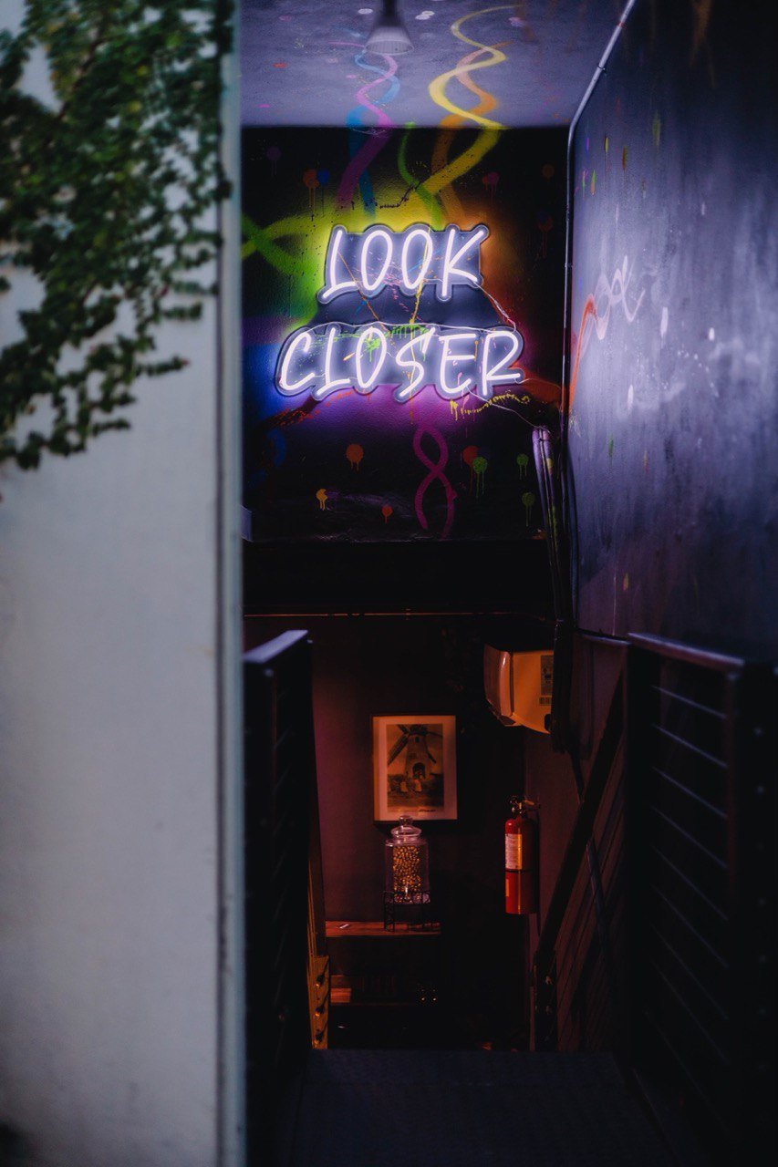 Neon sign that says 'LOOK CLOSER' at the top of a staircase in a dark interior space.
