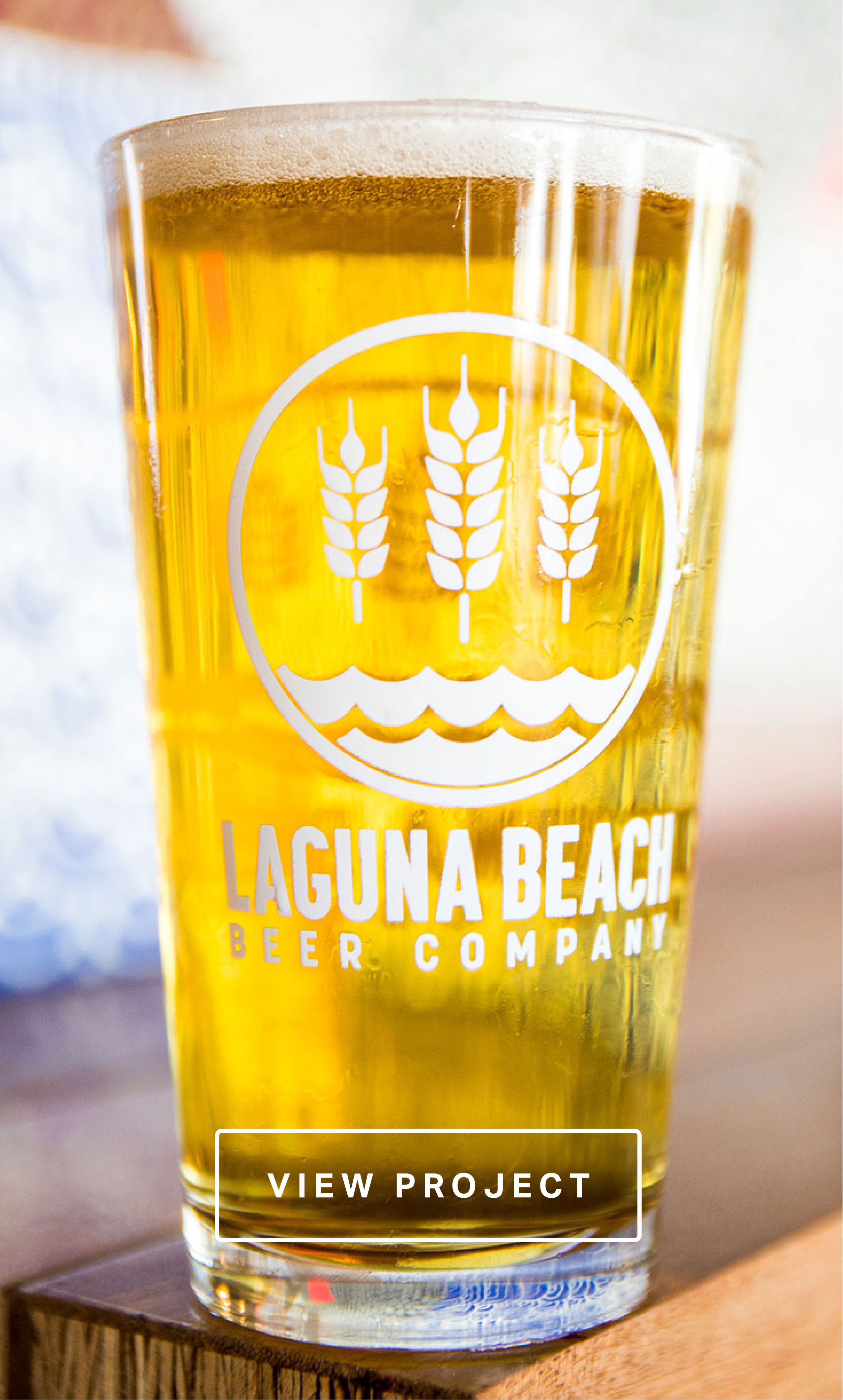 A glass of beer with the logo of Laguna Beach Beer Company, featuring three wheat stalks above waves, and a button labeled "View Project."