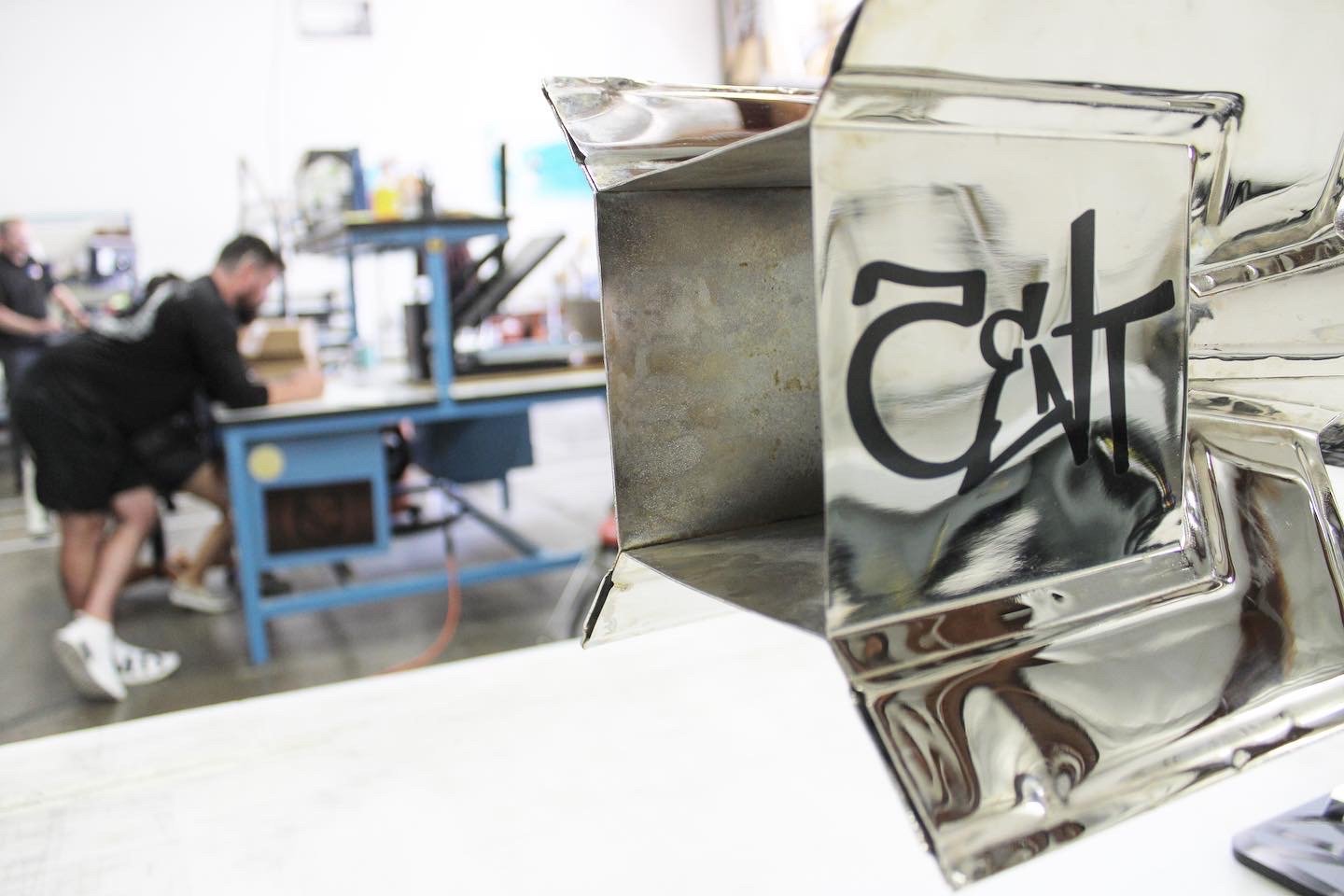 Close-up of a polished metal piece with a logo that reads 'Craft' reflected on its surface, with a blurred workshop background showing two people working at a blue workbench.