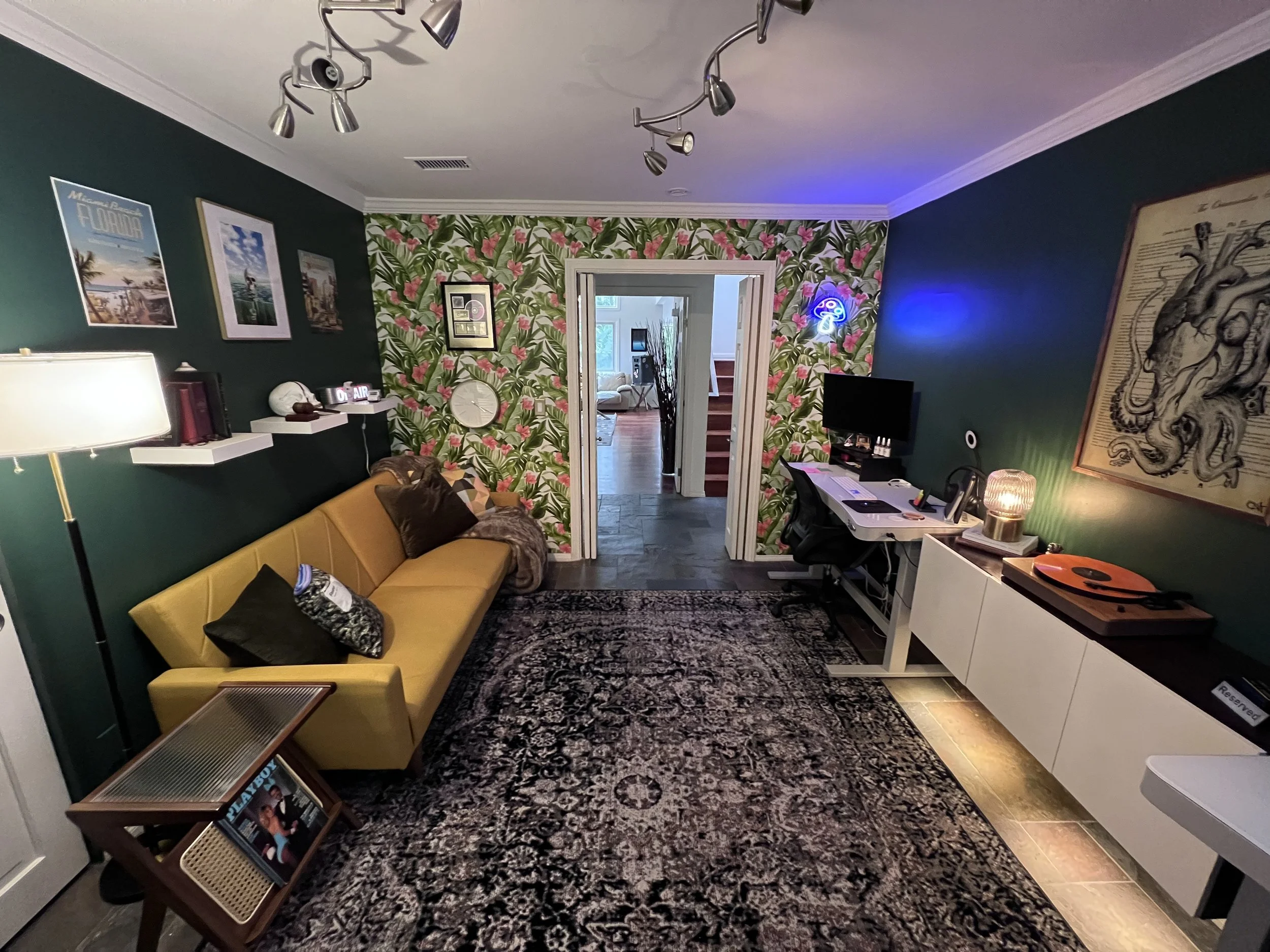A cozy living room with a yellow couch, a black patterned area rug, a white sideboard, a desk with a computer monitor, and tropical-themed wallpaper on one wall. There are framed pictures and a neon light on the walls, and a blue mushroom-shaped ligh