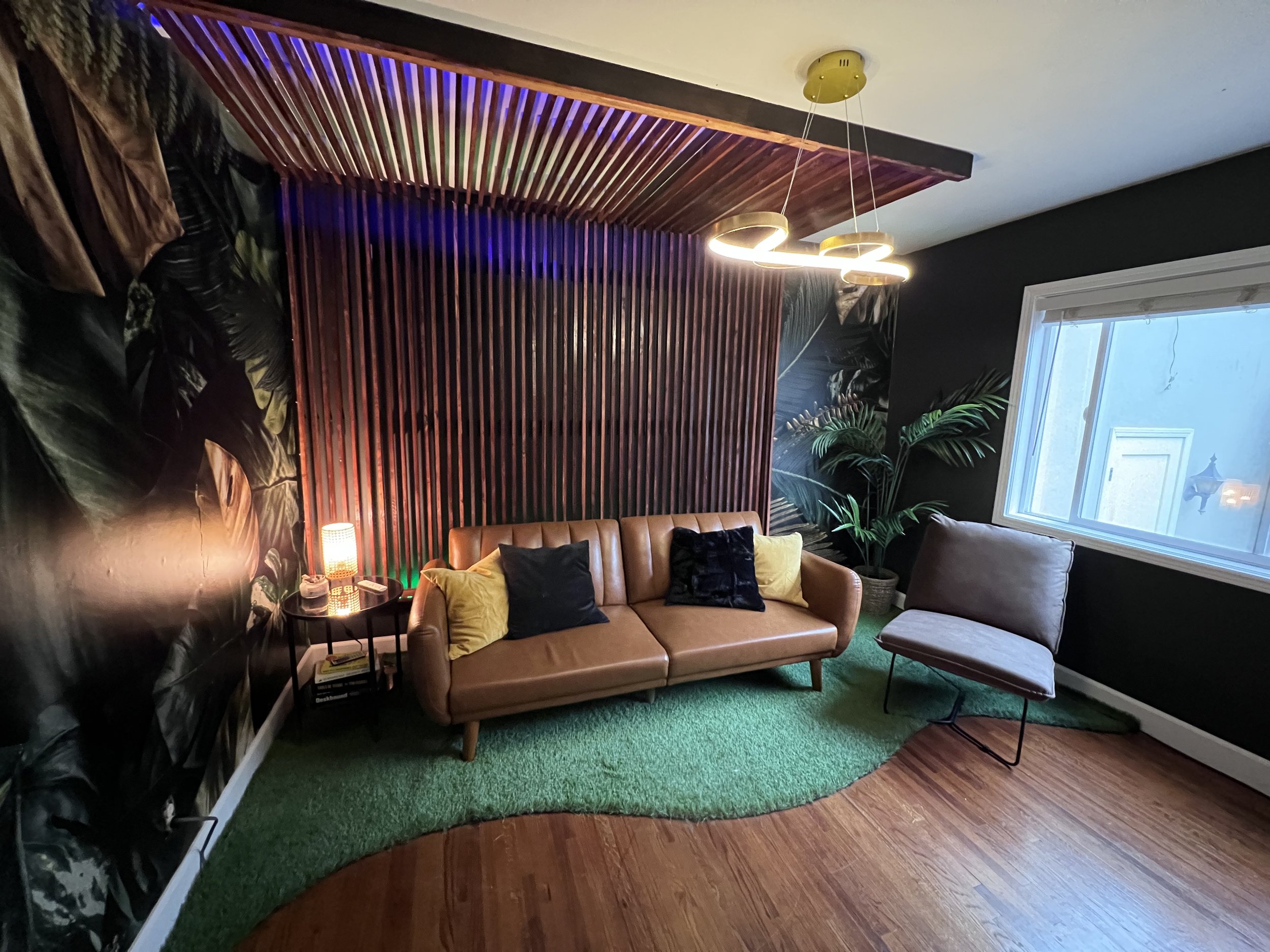 Living room with a tan leather sofa, a side table with a lamp, and a gray chair, decorated with pillows, a tropical plant, and wood wall accents, with a window on the right.