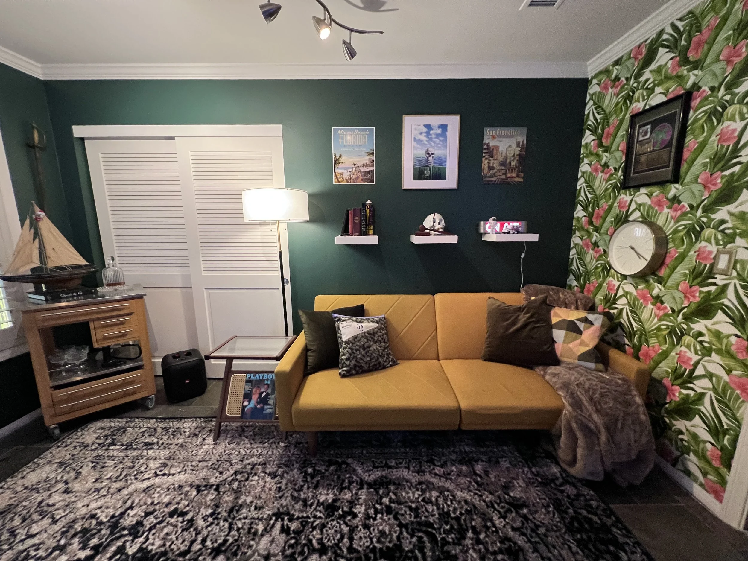 Living room with green walls, floral wallpaper accent wall, yellow sofa with pillows, patterned rug, wooden side table with shelves, white closet doors, floor lamp, wall art, and a small model boat on a piece of furniture.