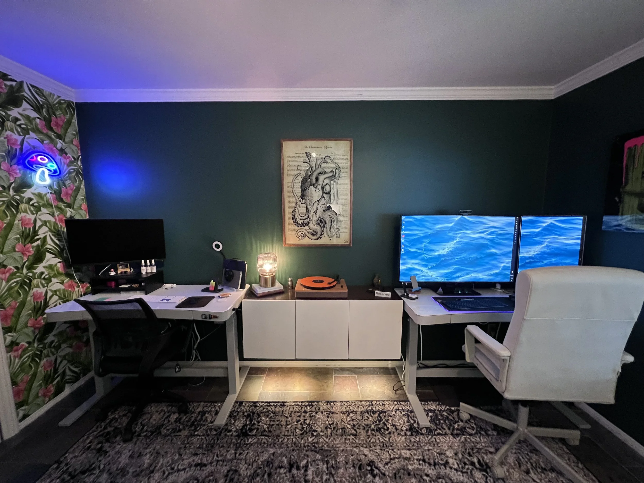 A home office with two desks, one with a computer monitor, and the other with a dual monitor setup. There's a floral wallpaper on the left wall with neon lighting, a framed black and white drawing of a dragon on the green wall, and various desk acces