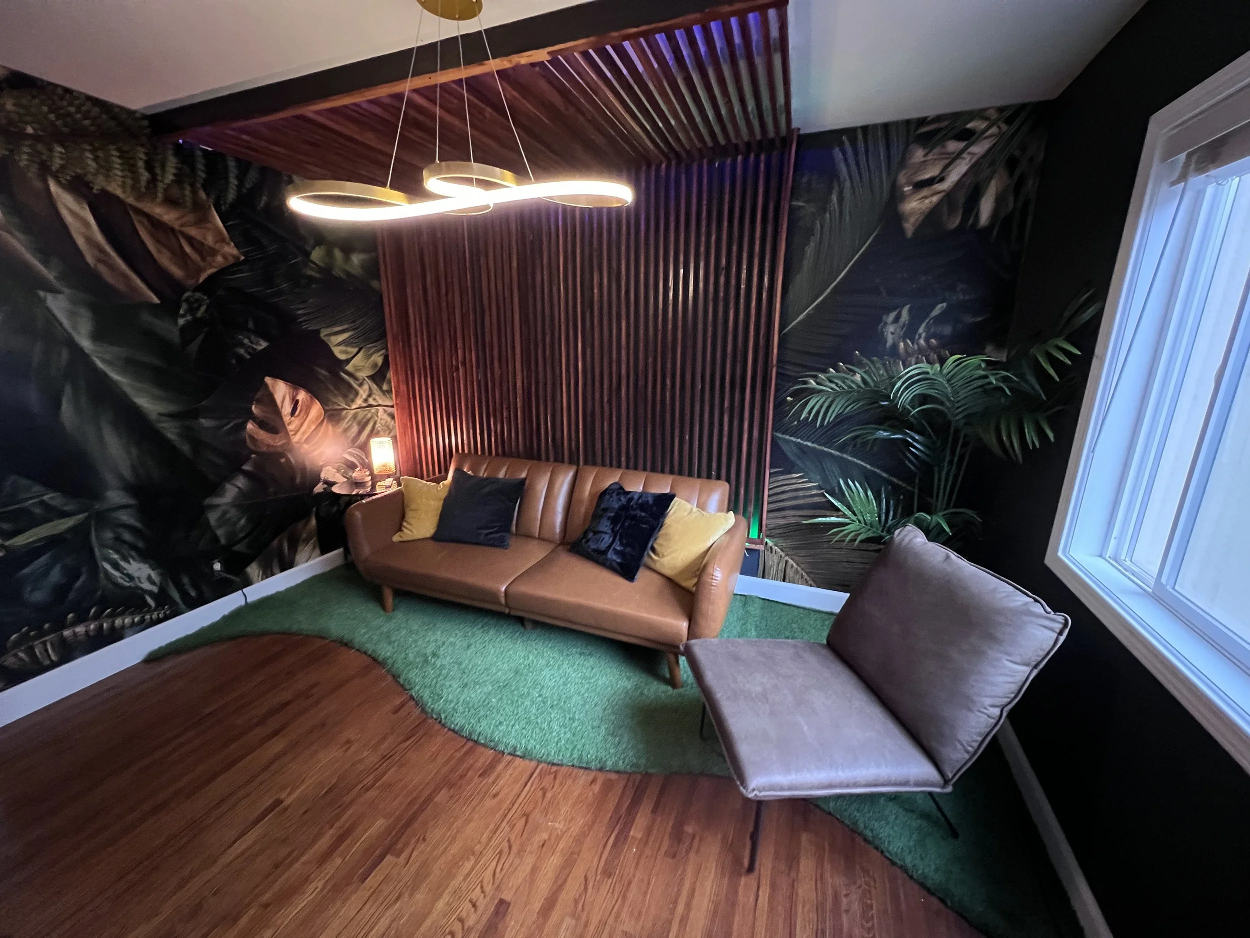 A cozy living room corner with a tan leather couch, decorative pillows, a modern lounge chair, a large window, a leafy plant, and a dark wall with tropical wallpaper, illuminated by stylish hanging lights.