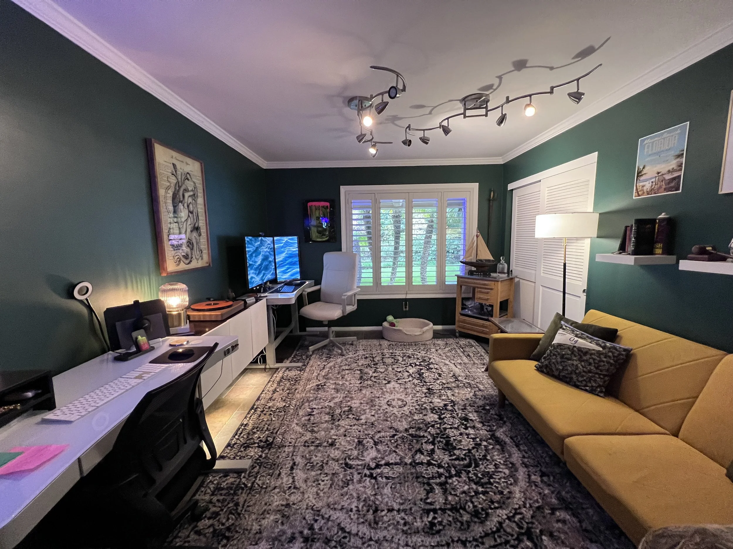 Living room with green walls, a yellow sofa with pillows, a white desk with a computer and black chair, a white armchair, a patterned rug, a window with shutters, a floor lamp, bookshelf, and ceiling lighting fixture.