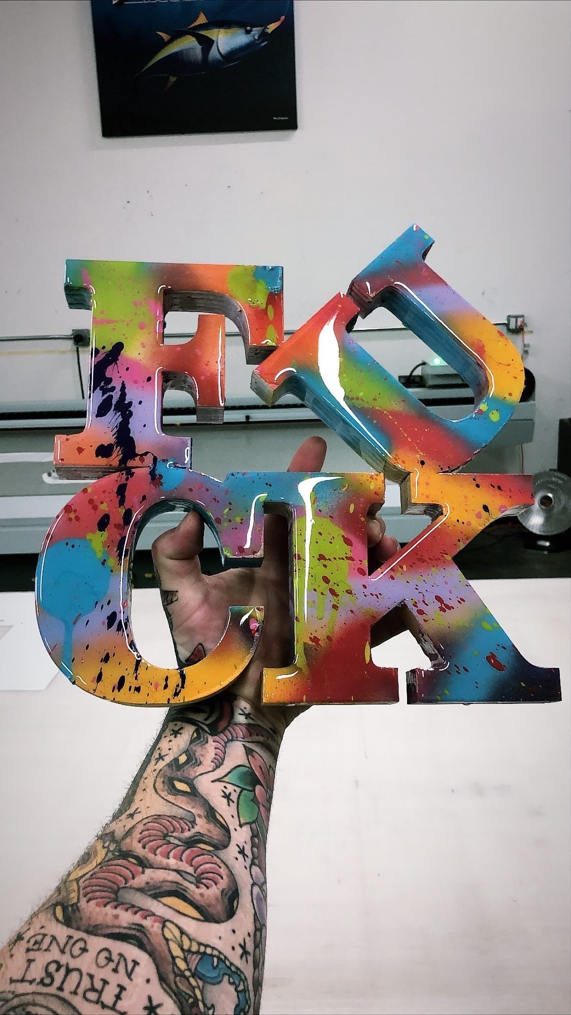 Colorful, splatter-painted, 3D-letter sign spelling "LOVE," being held by a tattooed hand against a plain wall, with a fish painting on the wall in the background.