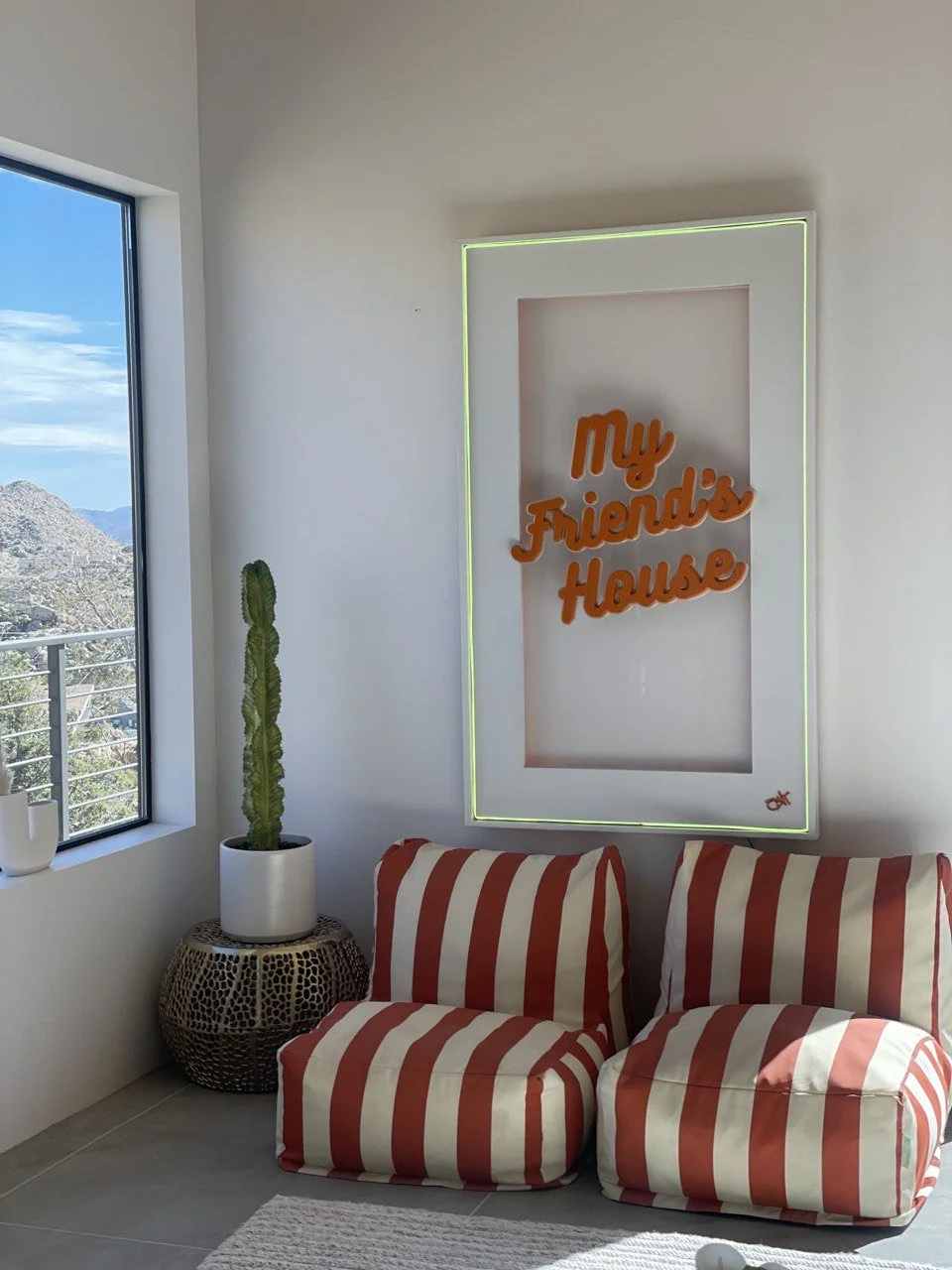 A cozy corner with a white wall, a large window showing mountains and blue sky, a cactus in a white pot on a patterned side table, and two red and white striped cushions on the floor. A neon sign reading 'My Friend's House' is mounted on the wall.