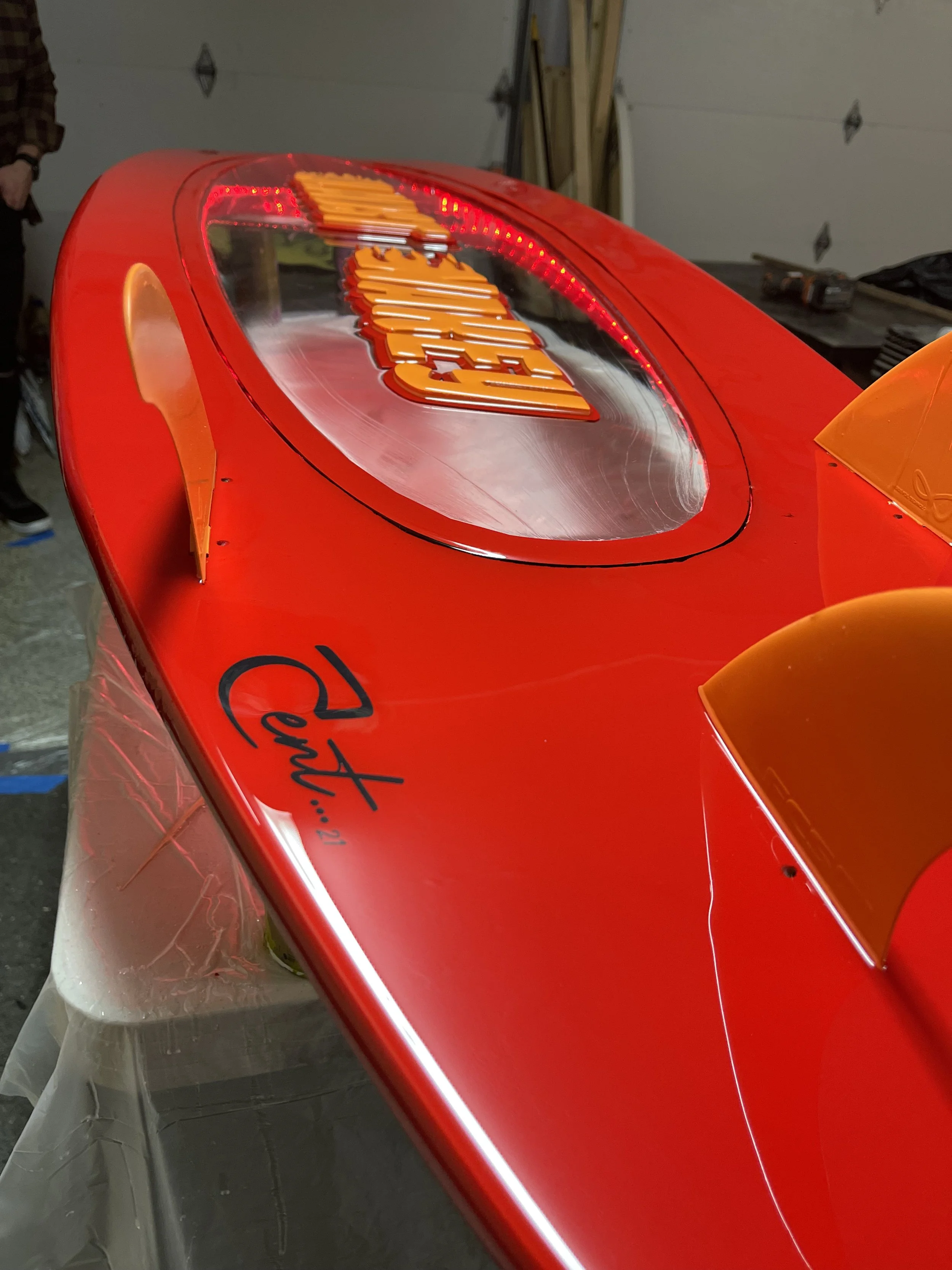 Close-up of a red race car with orange accents, signed by a driver, with the name 'Ayrton' visible near the edge and a large logo on the windshield.