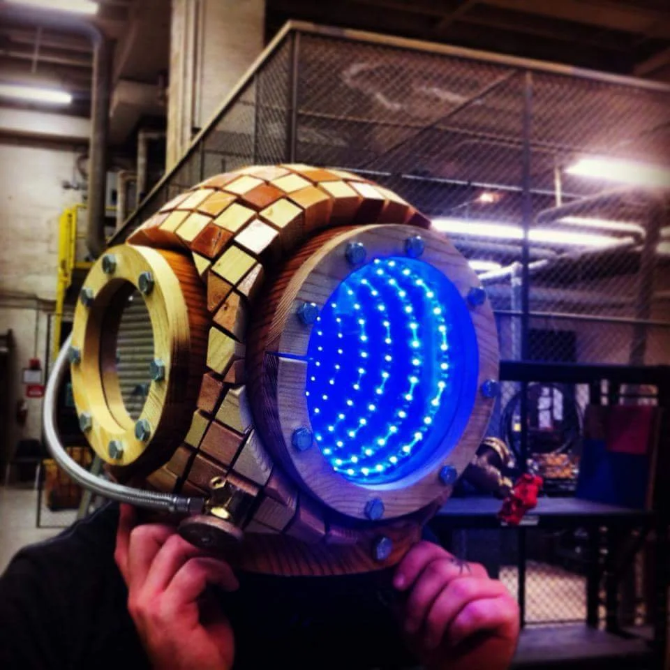 Person holding a large, steampunk-inspired, futuristic helmet with a blue LED light display, made of wood and metal, in an industrial workshop.