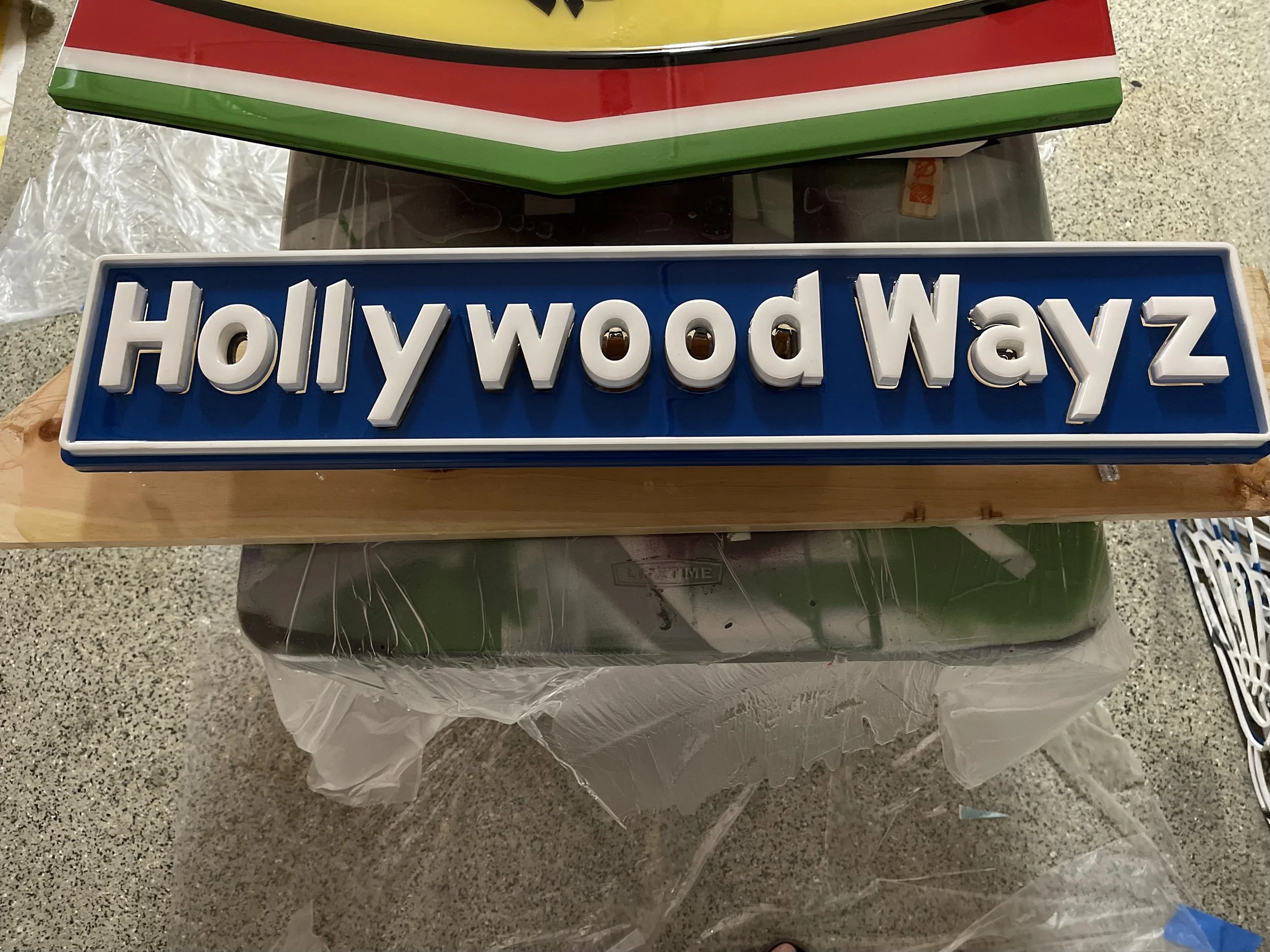 A street sign reads Hollywood Wayz with white letters on a blue background, mounted on a wooden board.