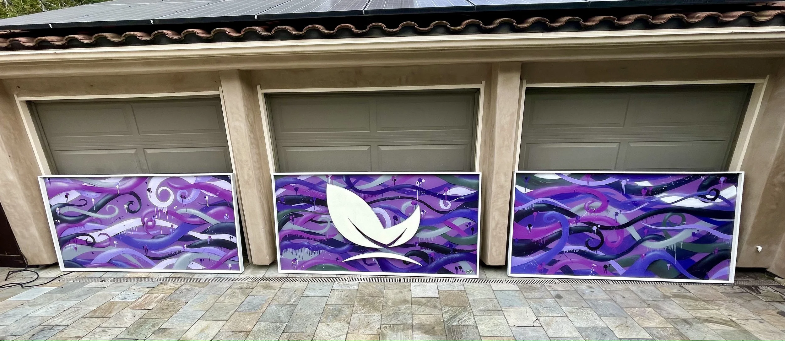 Three colorful paintings leaning against a beige garage with three closed garage doors. Each painting features abstract swirling purple, pink, and black patterns with white accents. The middle painting has a white leaf symbol in the center.