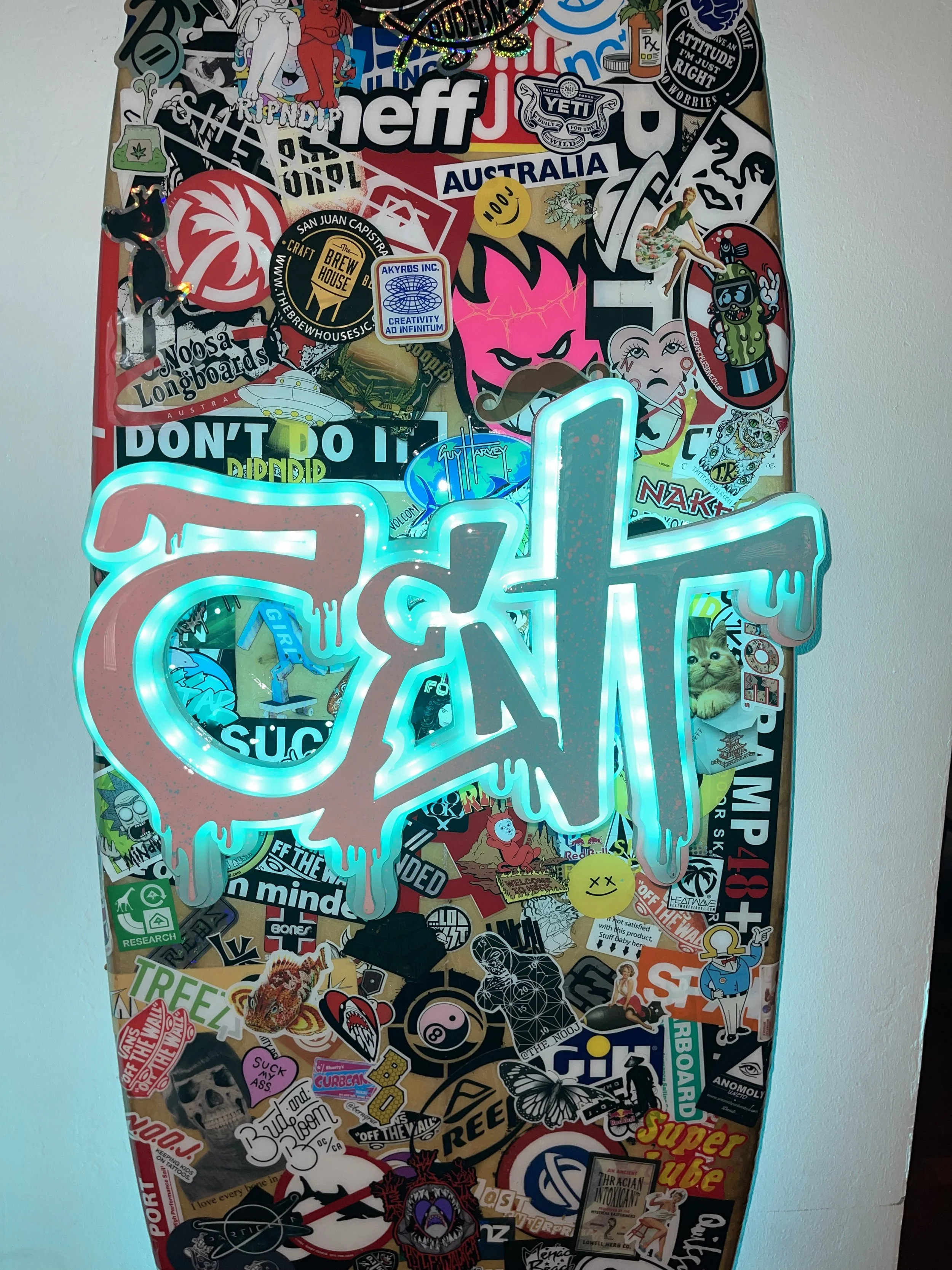 A skateboard deck decorated with numerous stickers and decals, featuring a neon sign that spells out a word in a dripping, graffiti style.