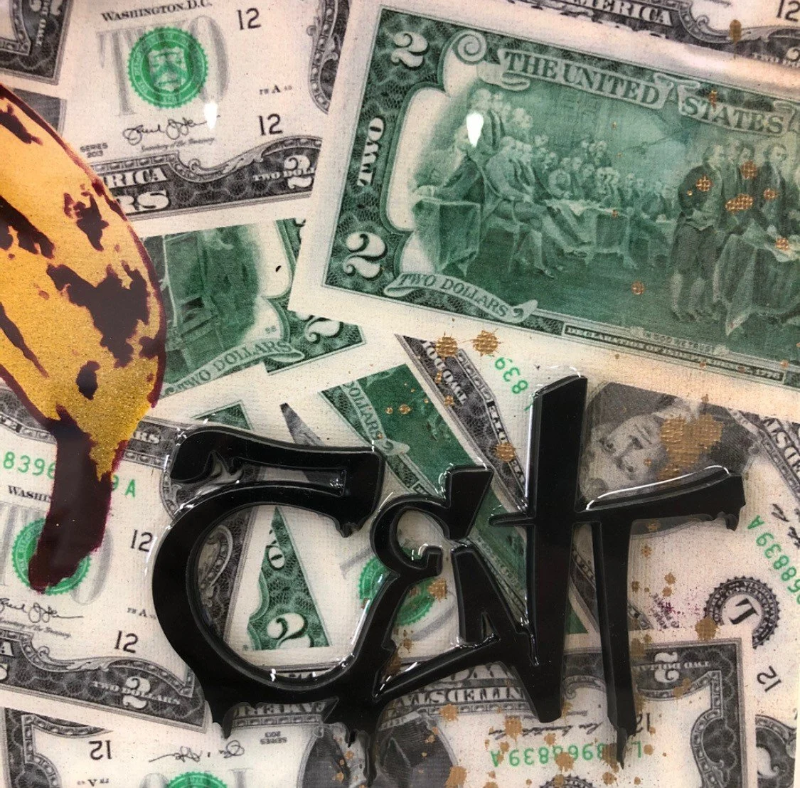 A collection of U.S. currency notes including a two-dollar bill, a twenty-dollar bill, and a one-dollar bill, with black decorative letters spelling 'eat' placed on top of the bills. There is also a piece of yellowish-orange fabric or clothing partia