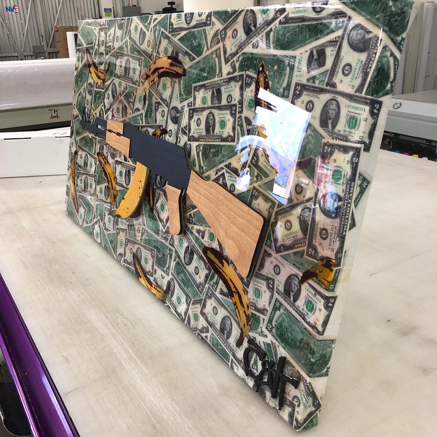 A decorative box with a dollar bill pattern and an image of a rifle on the front. The box is on a flat surface in a workshop or warehouse with other equipment in the background.