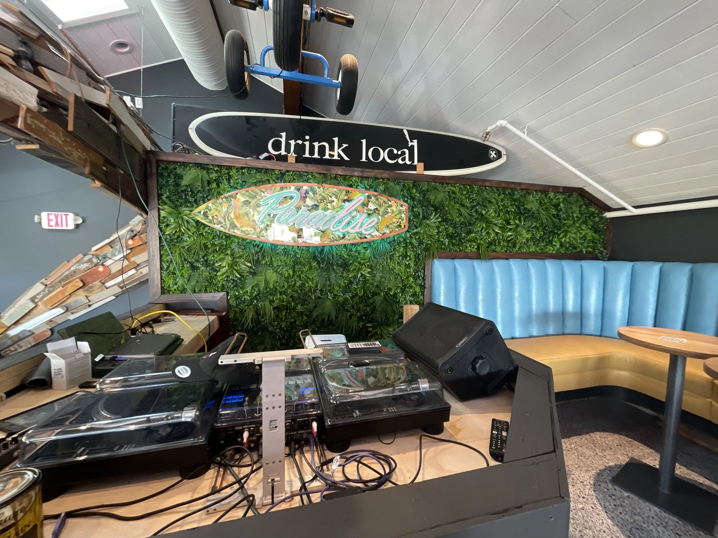 Interior of a bar or lounge area with DJ equipment, a green leafy wall with a colorful neon sign that reads 