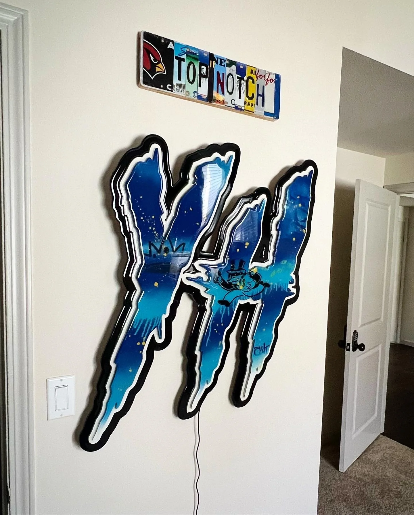 Decorative wall art of the Phoenix Suns logo with a graffiti-style design, blue and white colors, and a cartoon character in the center, mounted on a white wall.