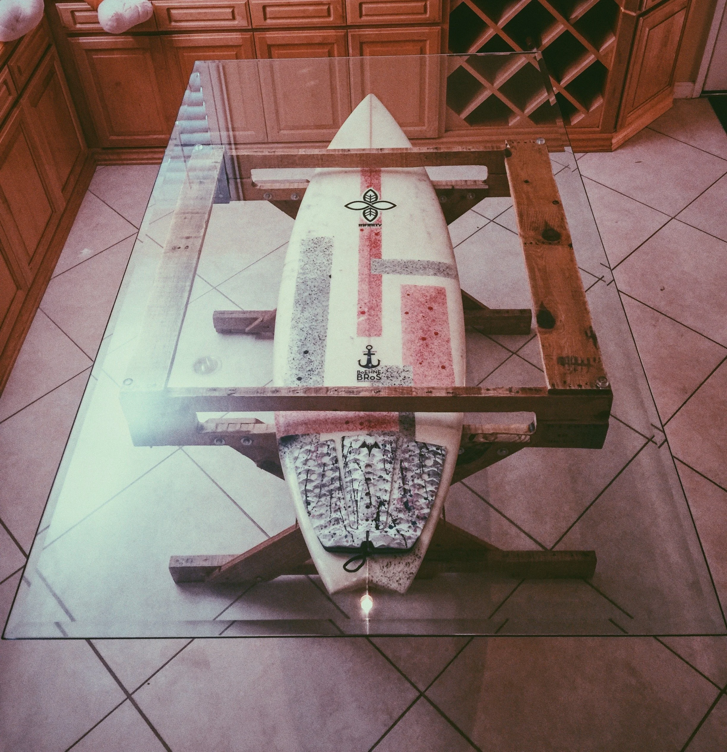 A surfboard inside a glass-top table in a kitchen with wooden cabinets and tiled floor.