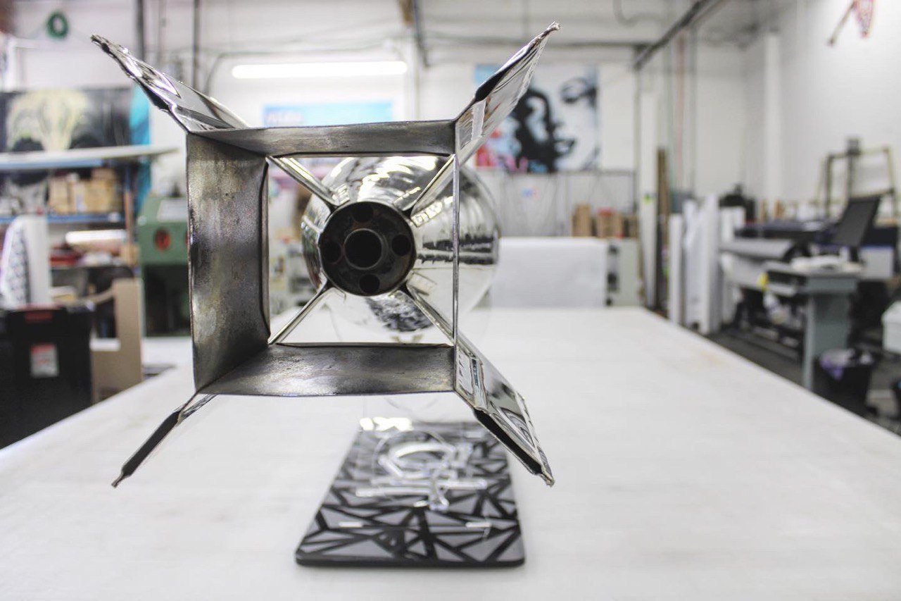 Close-up of a metallic rocket model mounted on a patterned stand, with an industrial workshop in the background.