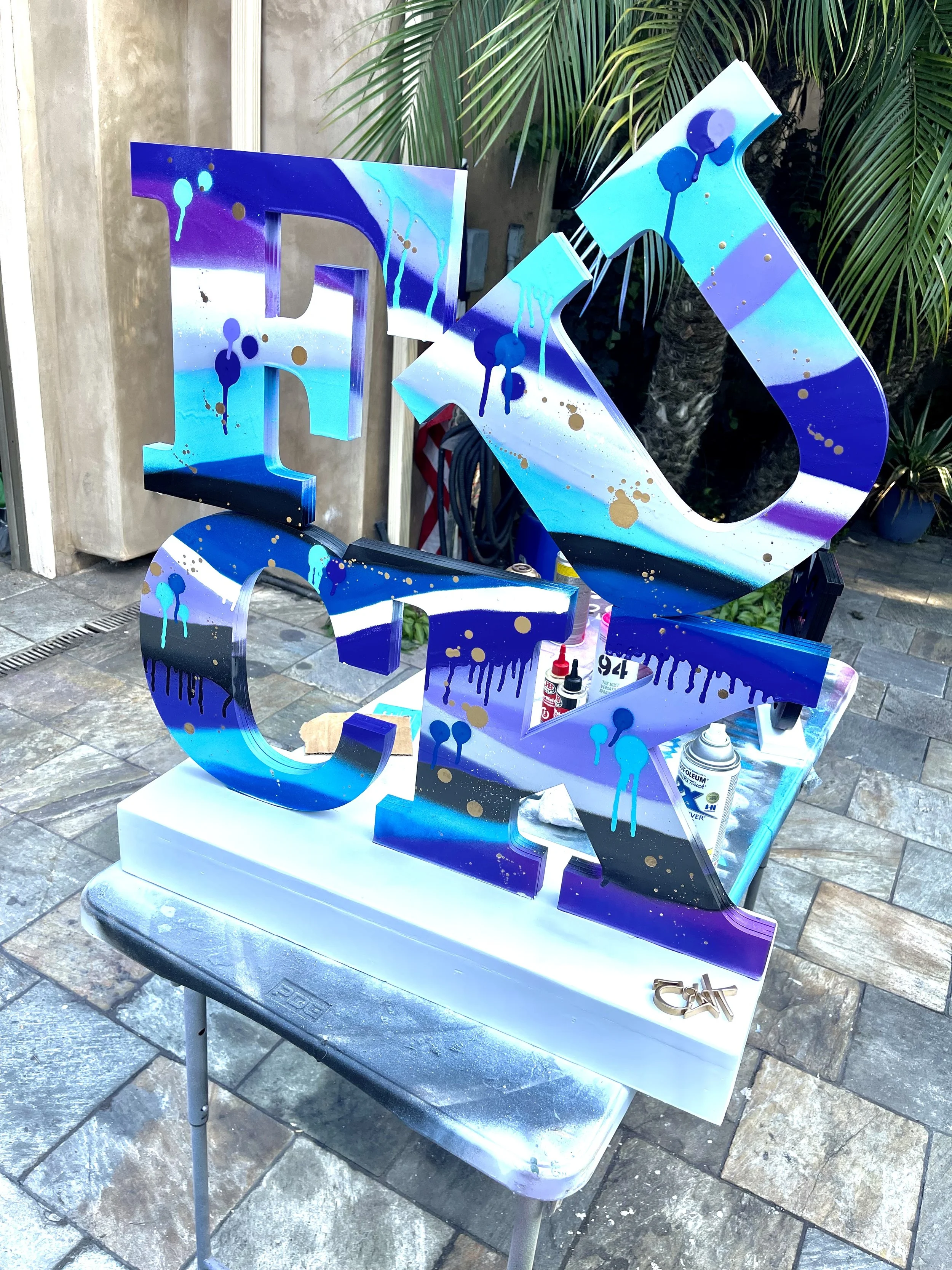 Colorful painted metal letters spelling 'LOVE' on a white surface, placed outdoors with spray paint supplies and tools around.