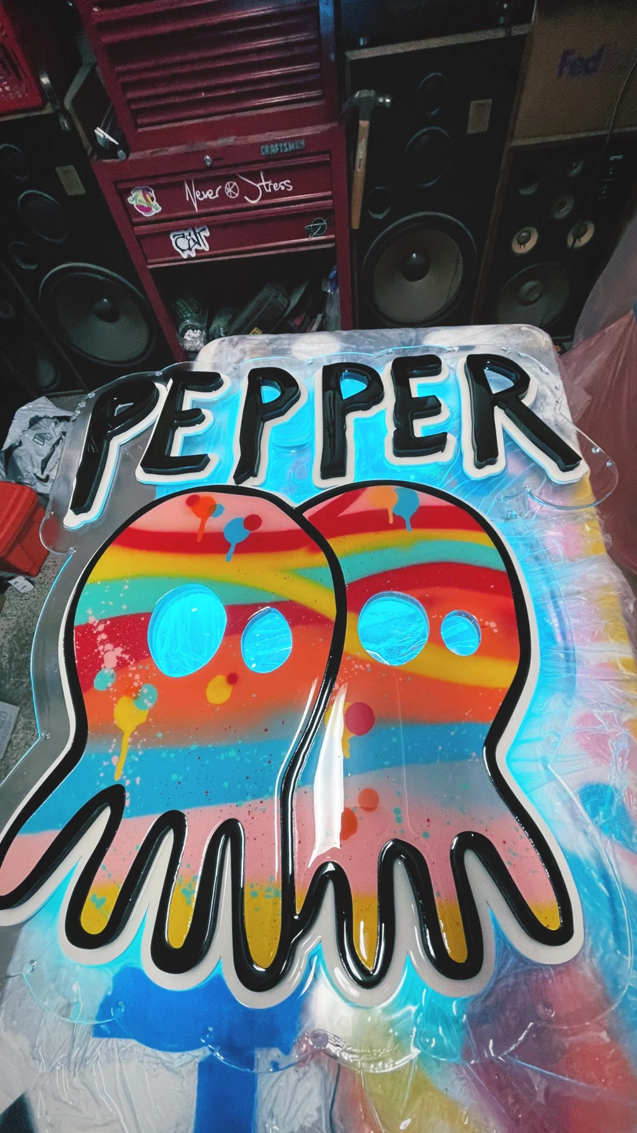 A colorful, illuminated sign featuring the phrase "PEPPER" above two stylized pepper shapes with a rainbow-colored splatter pattern, set against a workshop background with tools and speakers.