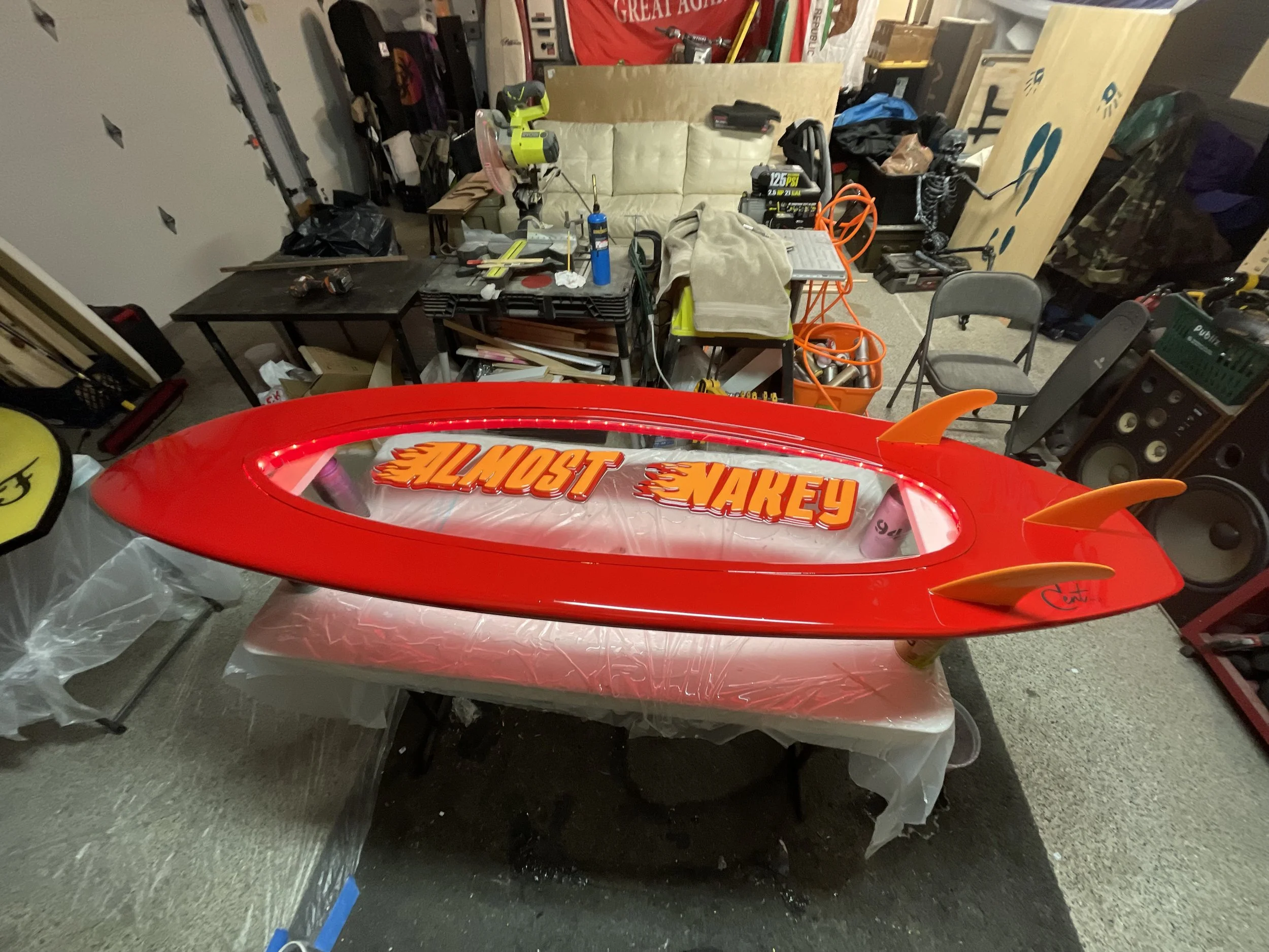 A red remote-controlled speedboat with orange fins, named 'Naked' and 'Most', resting on a plastic-covered table in a cluttered workshop or garage.
