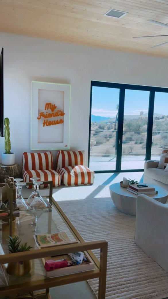 Living room with striped orange and white armchairs, a white sofa, glass coffee table, indoor plants, a large window with a view of the outdoors, and a wall sign that says 'My Friend's House'.