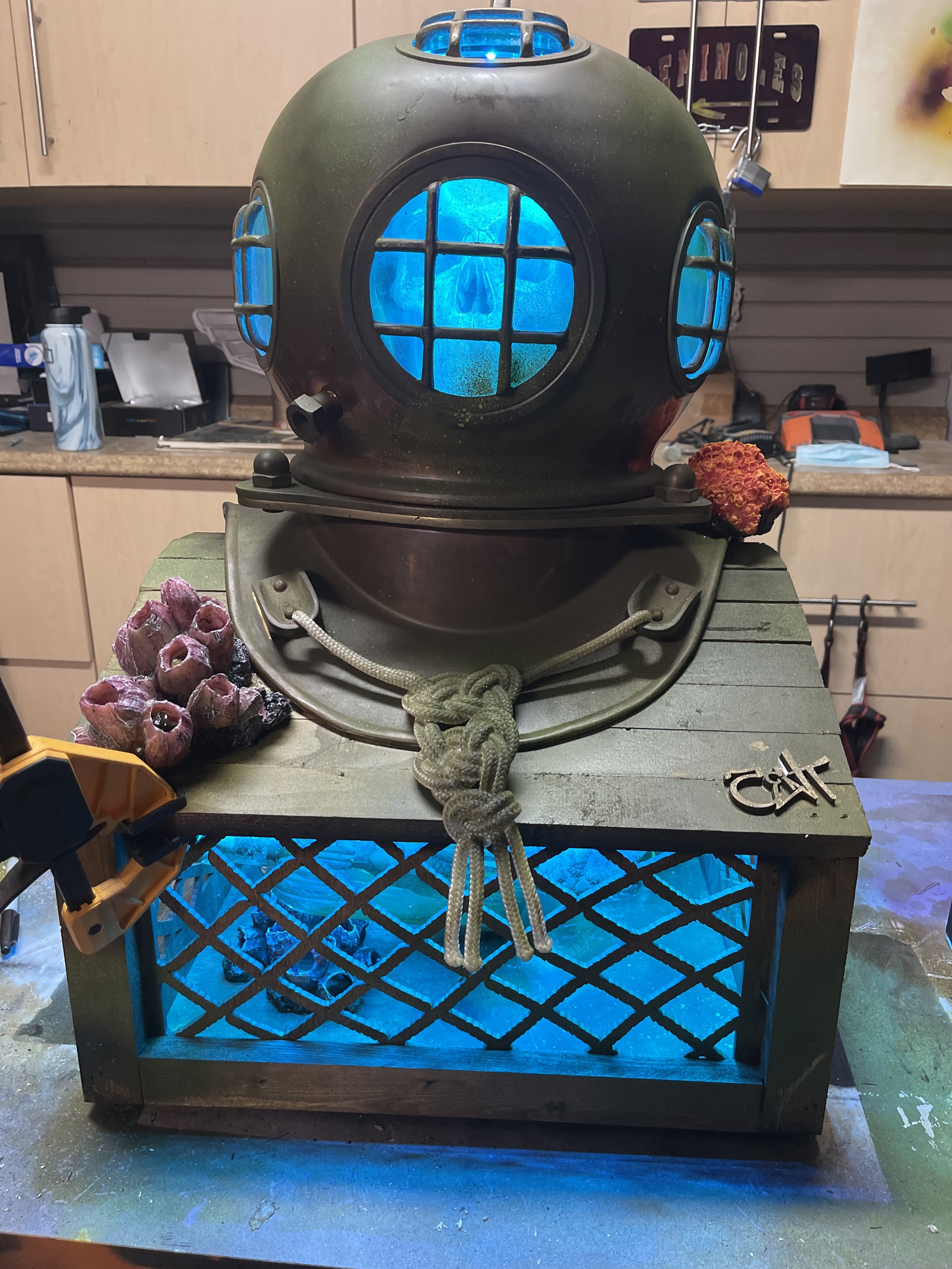 A vintage diving helmet with glowing blue windows placed on a wooden table. The table has a blue light underneath and a knot tied in a rope hanging from the front. There are decorative coral pieces and a metal tag with the word 'CAP' on it beside the