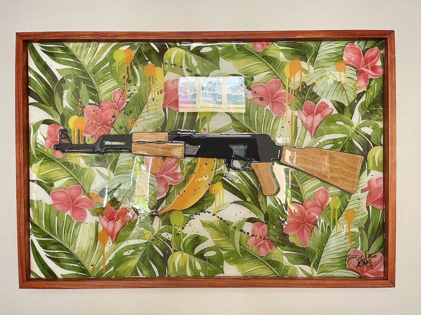 Framed artwork of a rifle with wooden parts and black metal, set against a background of green tropical leaves and pink flowers with yellow accents, with yellow paint splatters.