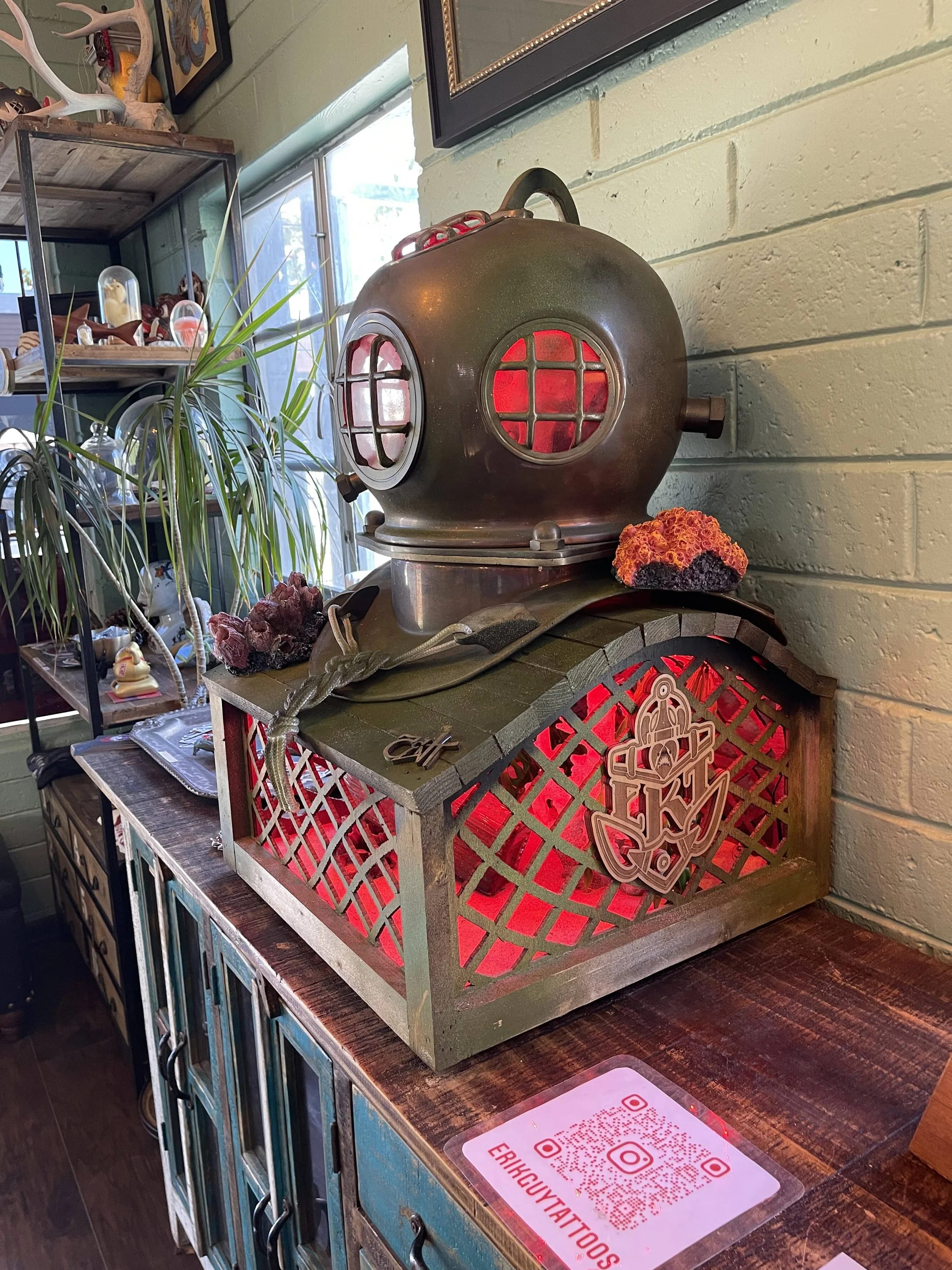 Decorative underwater-themed lamp resembling a vintage deep-sea diver helmet, placed on a wooden sideboard with a green lattice box and a coral rock, surrounded by plants and other decor items inside a room.