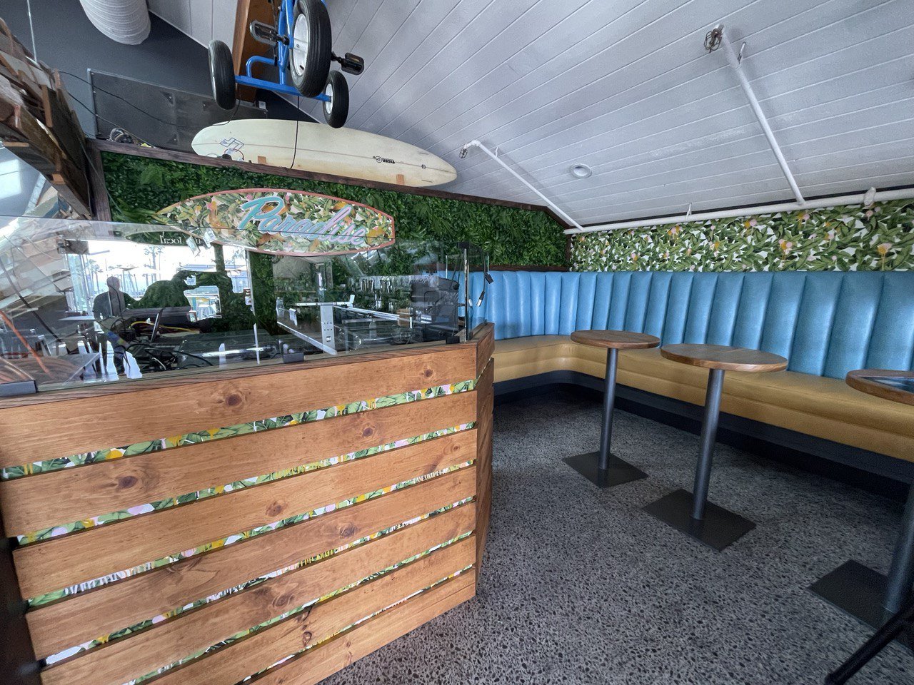 Inside a tropical-themed cafe with blue and yellow upholstered booth seating, small round wooden tables, a decorative green leafy wall, a surfboard, and a display case with a colorful sign that reads 'Palmeta'.