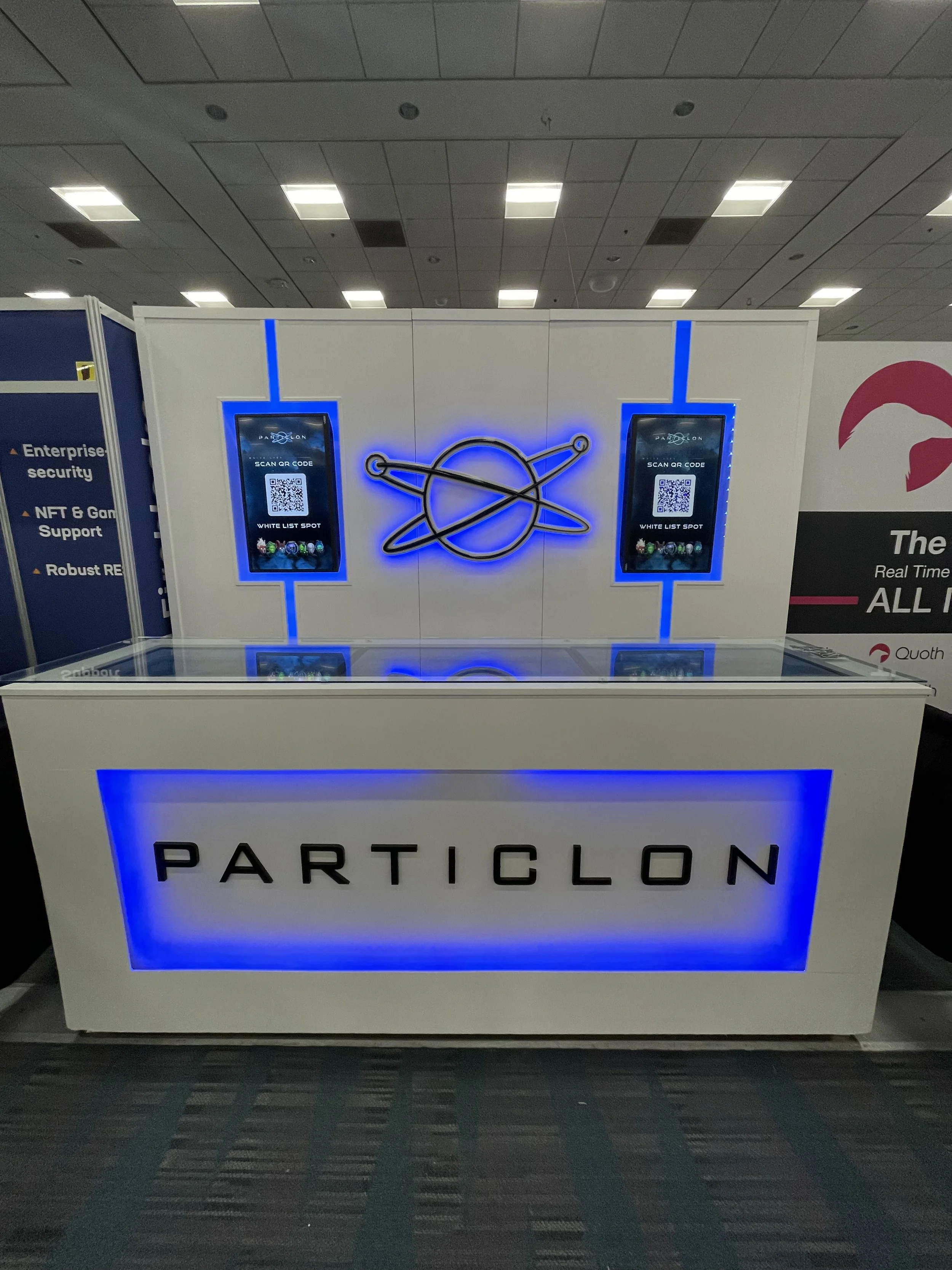 Trade show booth with a white counter displaying the word 'PARTICLON' in black letters and a space-themed logo on the wall behind it illuminated with blue neon lights, featuring two digital screens displaying QR codes.