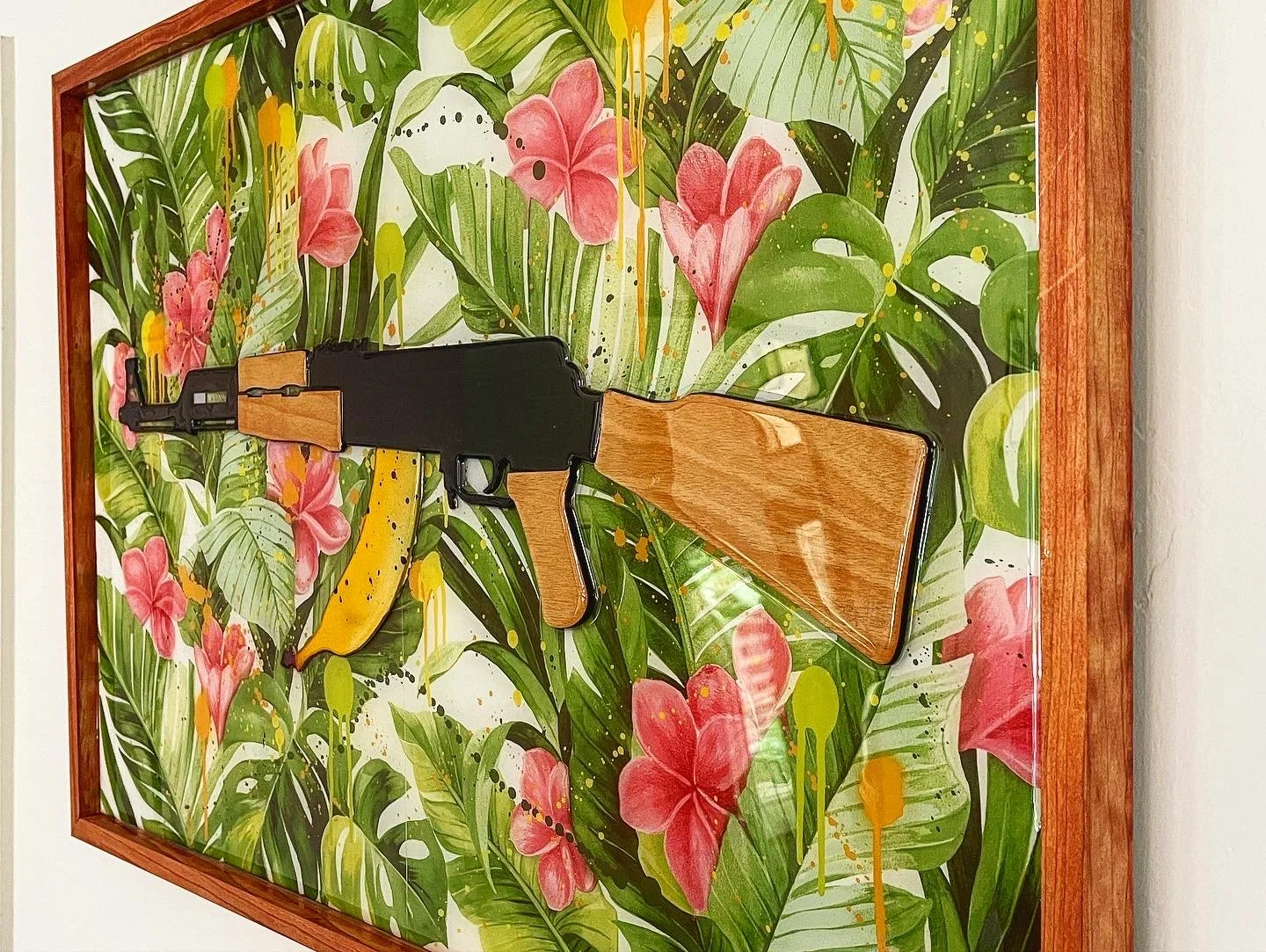 A framed artwork featuring a tropical leaf and flower background with a three-dimensional gun and banana sculpture mounted on it.