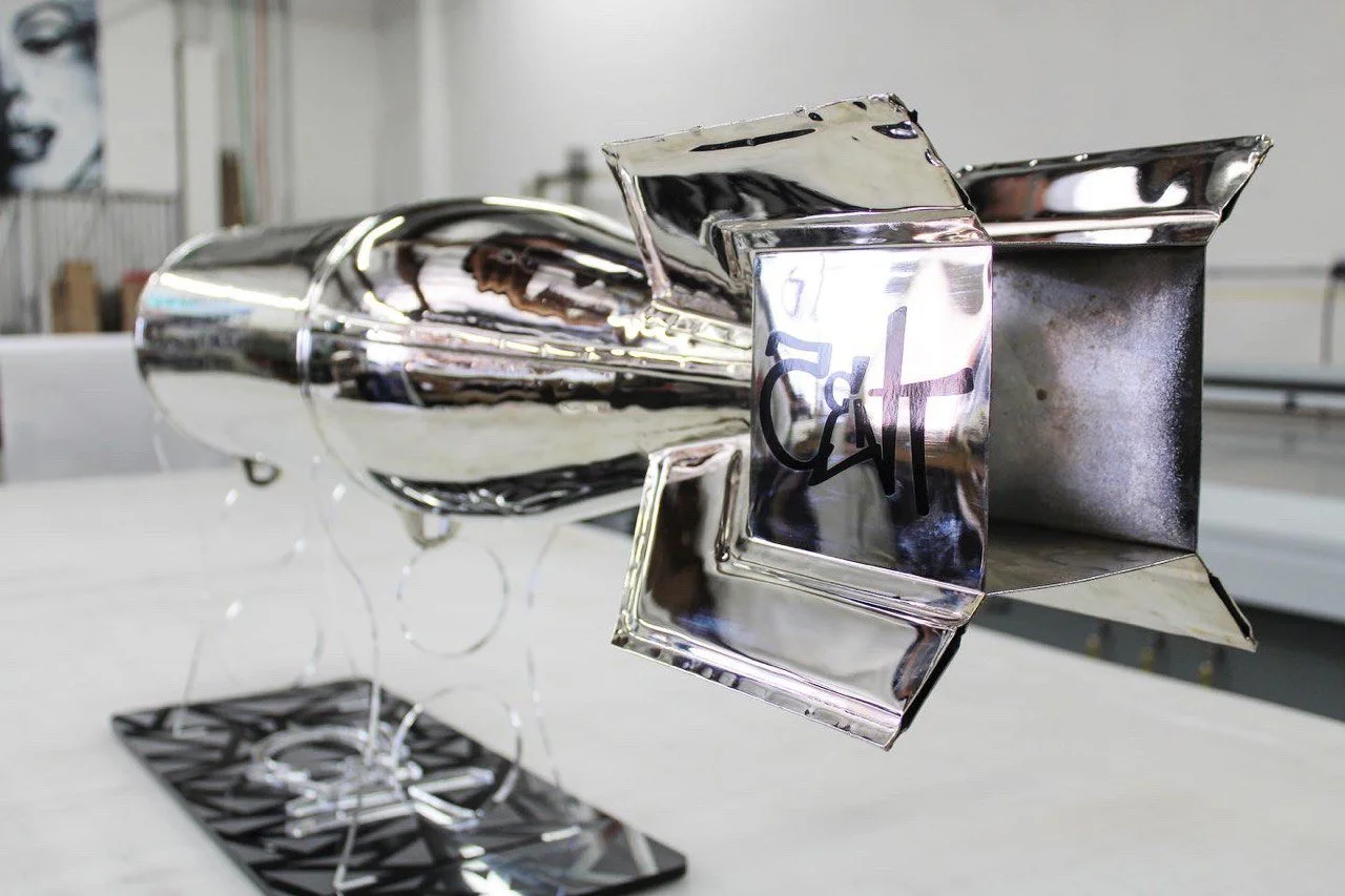A shiny metal rocket sculpture mounted on a black stand in a workshop or studio.