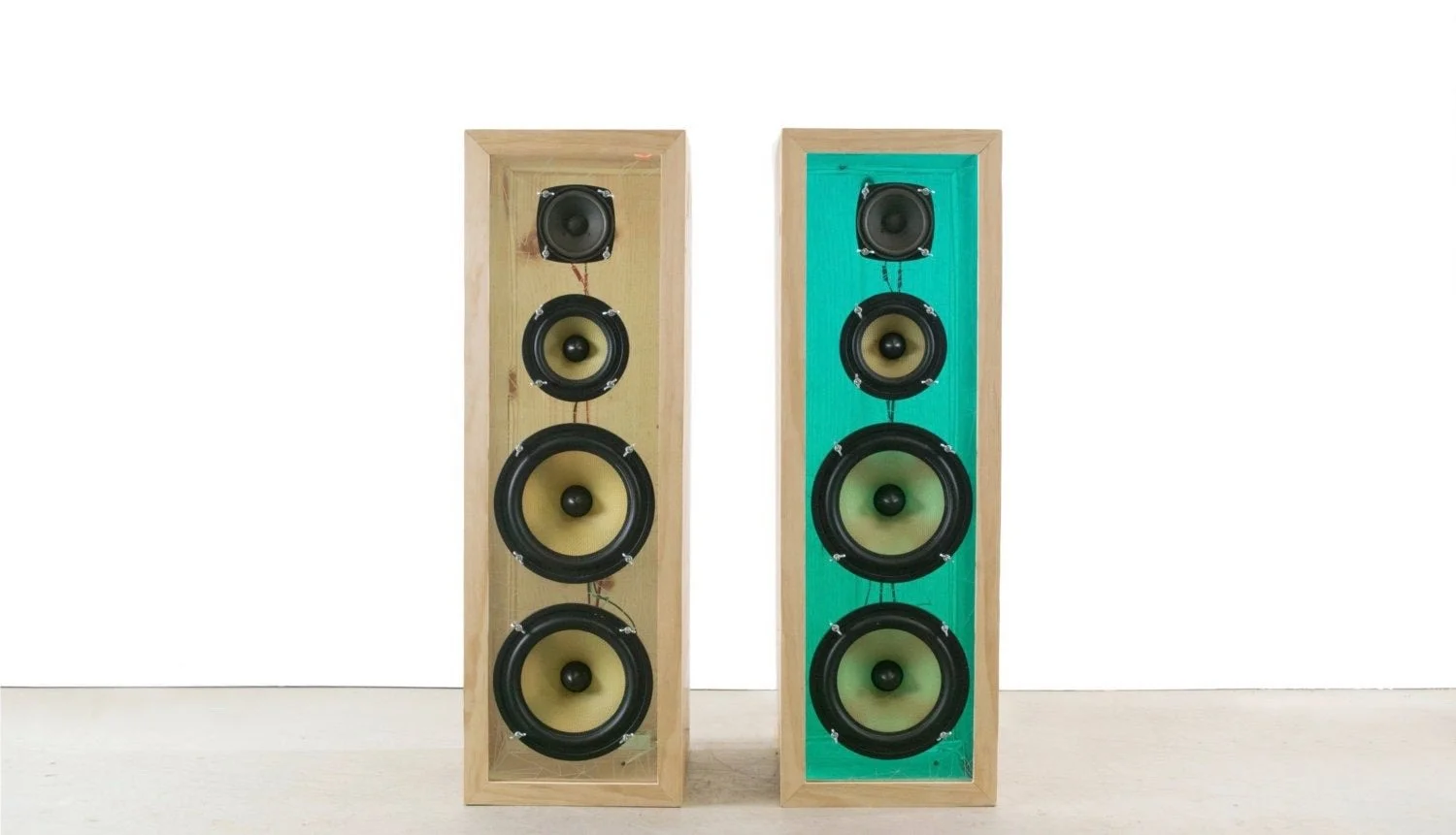 Two tall wooden speakers with multiple black and yellow speaker cones, one with a turquoise background panel, placed on a light table against a plain white wall.