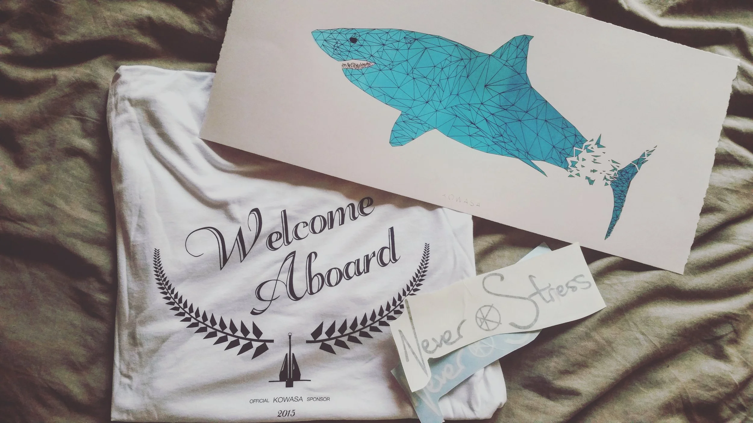 A white fabric banner with black text that says 'Welcome Aboard' and features a laurel wreath and a ship logo, along with a small handwritten note that says 'Never Stress'; a poster of a blue geometric shark with a fragmented tail.