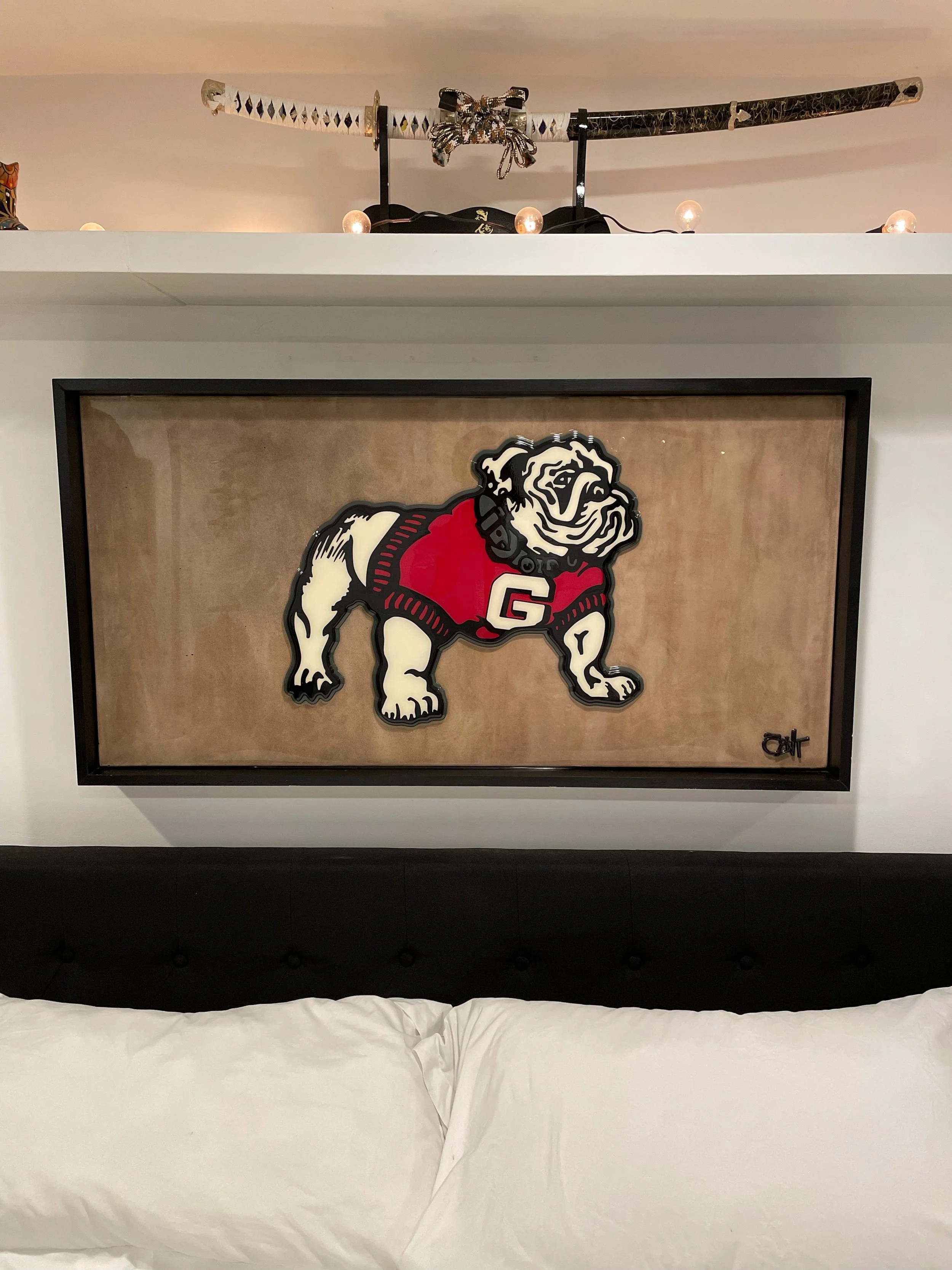 Decorative framed picture of a bulldog wearing a red football jersey with the letter G, hanging above a black headboard of a bed with white pillows.