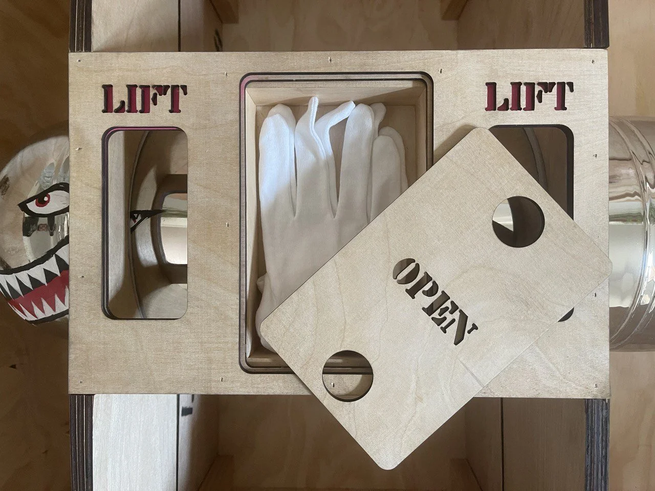 A wooden box with the words "LIFT" engraved on it, containing white gloves. There are two chipped wooden pieces with "LIFT" and a circular hole on each, placed on top of a glass of water. A paper mask with a shark face design is partly visible to the