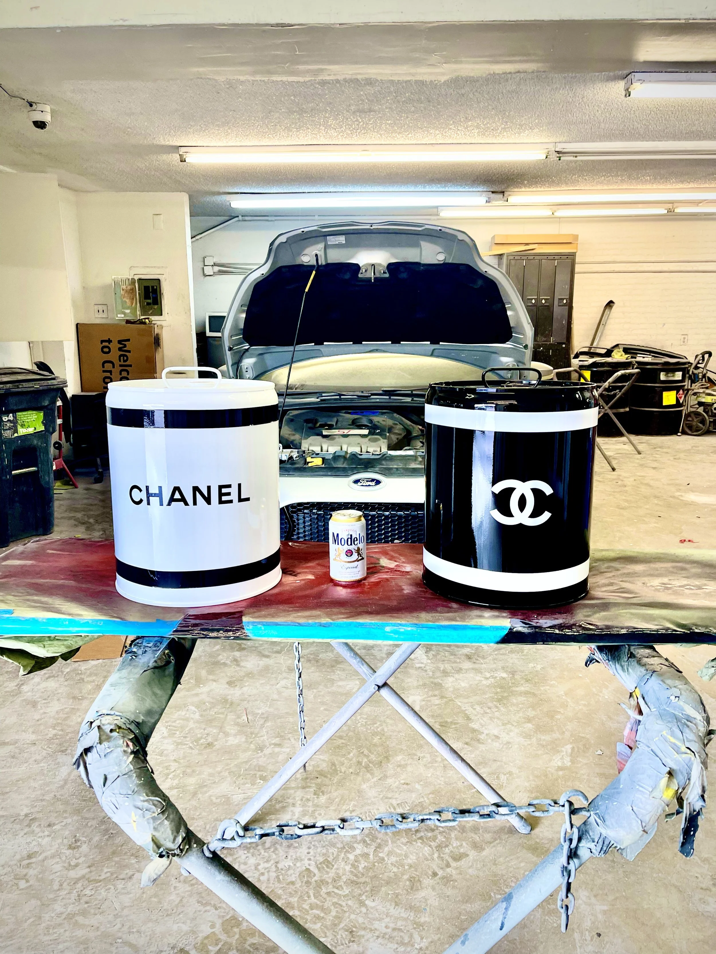 Two large Chanel and Chanel-inspired containers, a Modelo beer can, and a car with its hood open in a garage.