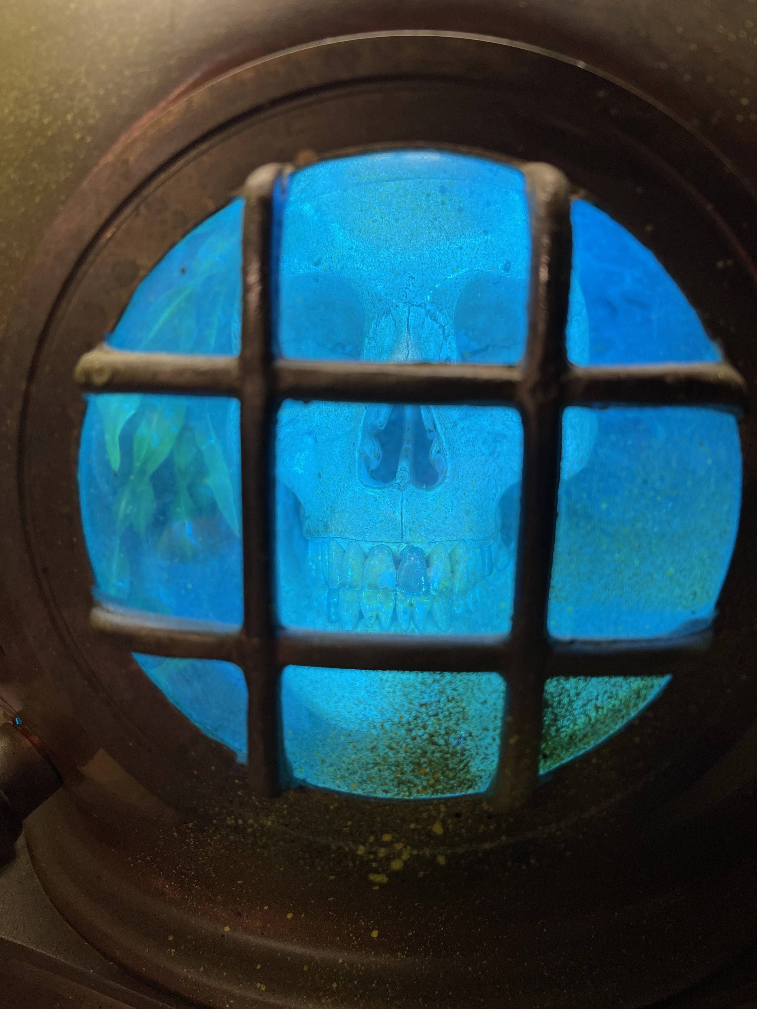 A skull is visible behind a barred, round window with a blue tint, inside a dark metallic frame, giving a spooky or eerie effect.