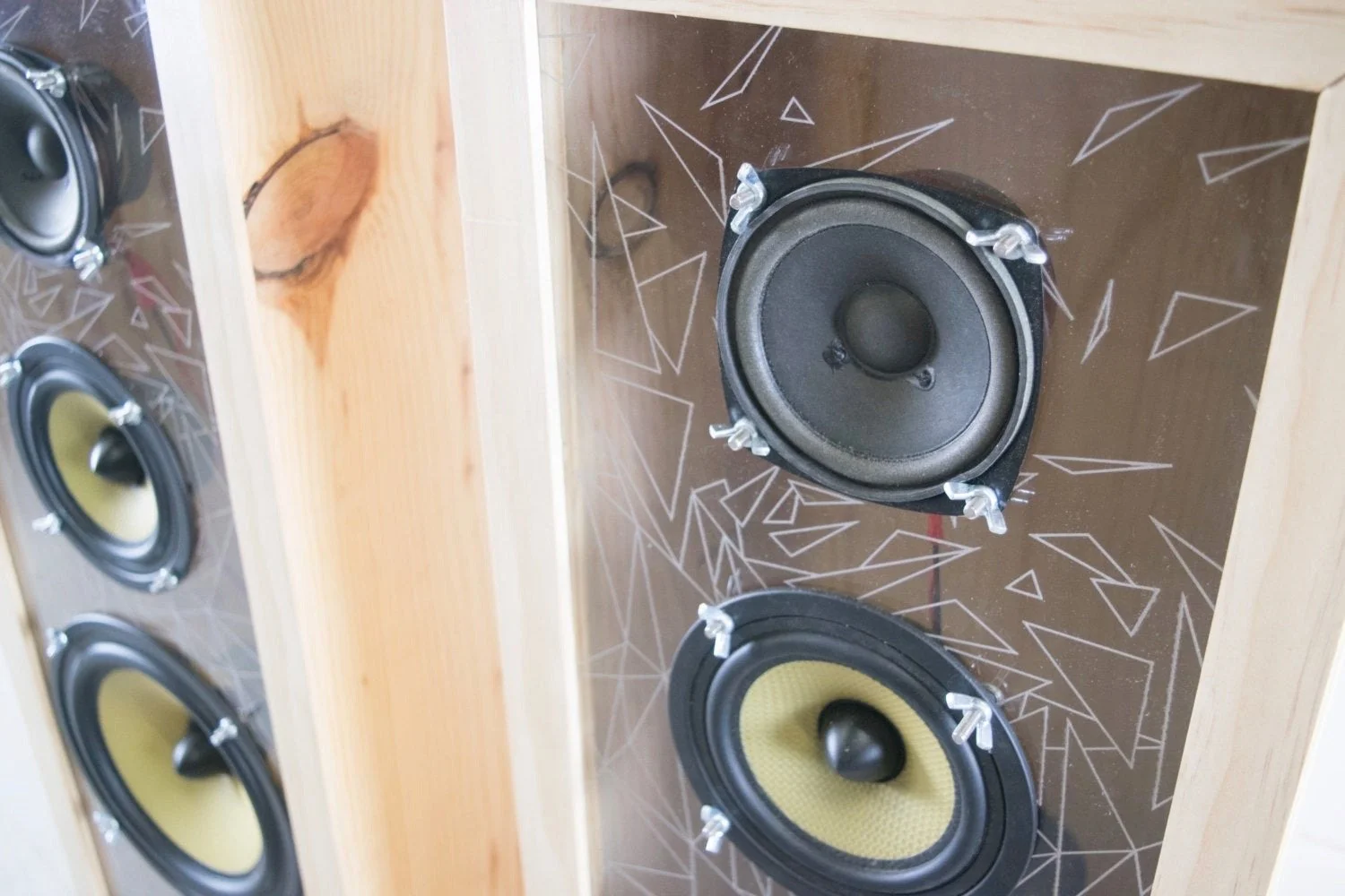 Close-up of a wooden speaker box containing four yellow and black speakers mounted on a brown back panel with white geometric line patterns.