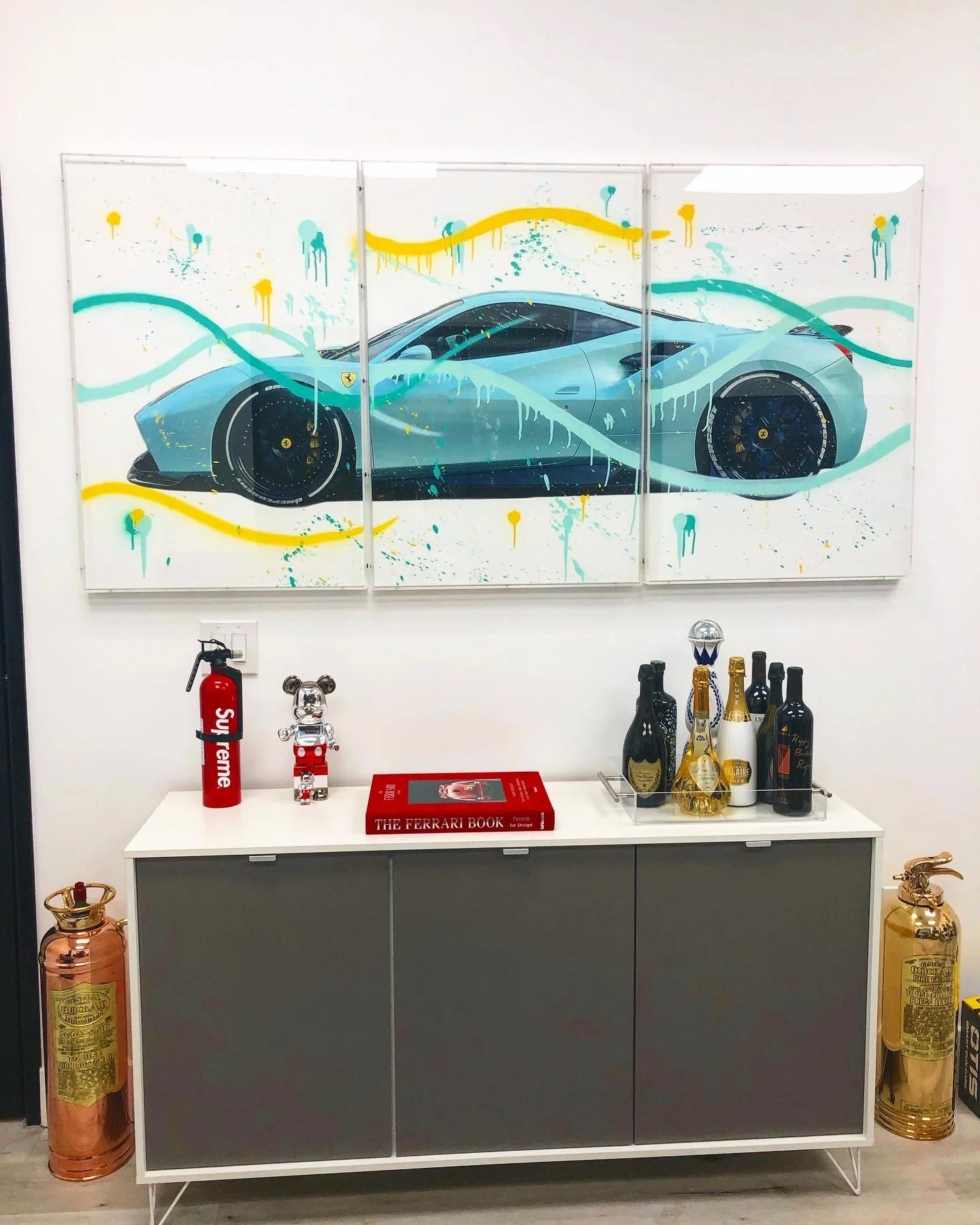 A modern white cabinet with a black and gray door, decorated with a red book titled 'The Ferrari Book,' a red Supreme fire extinguisher, a small silver bear figurine, and an assortment of wine bottles and champagne on a tray. Above the cabinet is a l