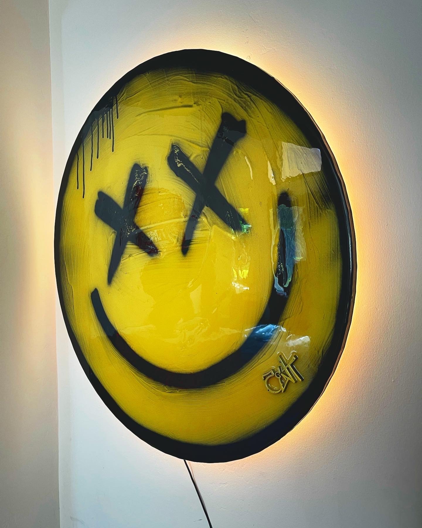 A round yellow wall clock with a black smiley face design and black graffiti-like markings, backlit with a halo of light on the wall.