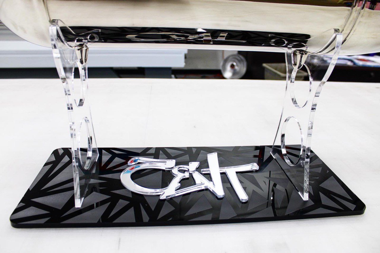 A black reflective base with a geometric pattern, supporting a clear acrylic stand, and a metallic decoration of the word 'CAT' on the surface.