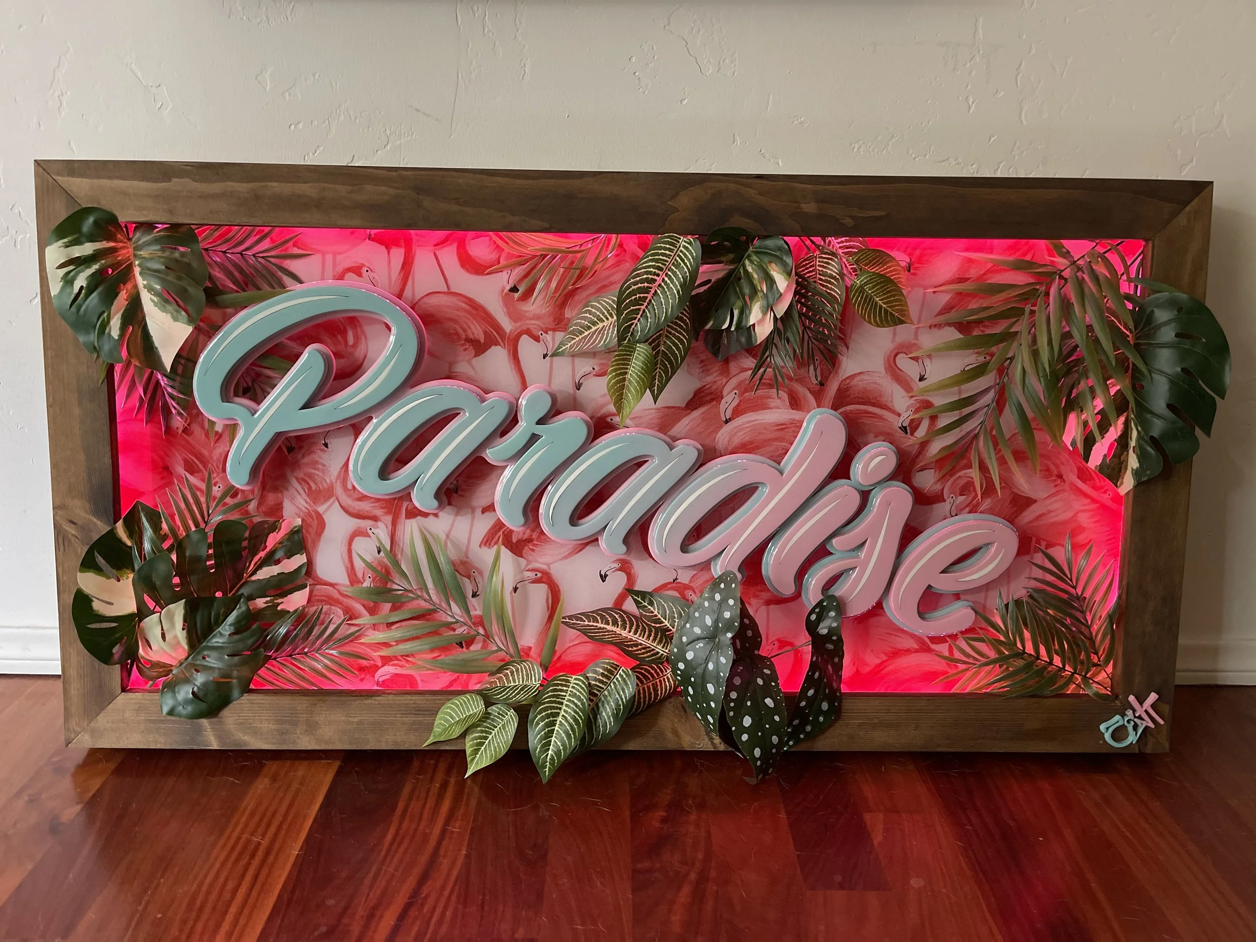 Decorative framed sign with the word 'Paradise' in light blue script, surrounded by tropical leaves and pink flamingo illustrations, with pink backlight.