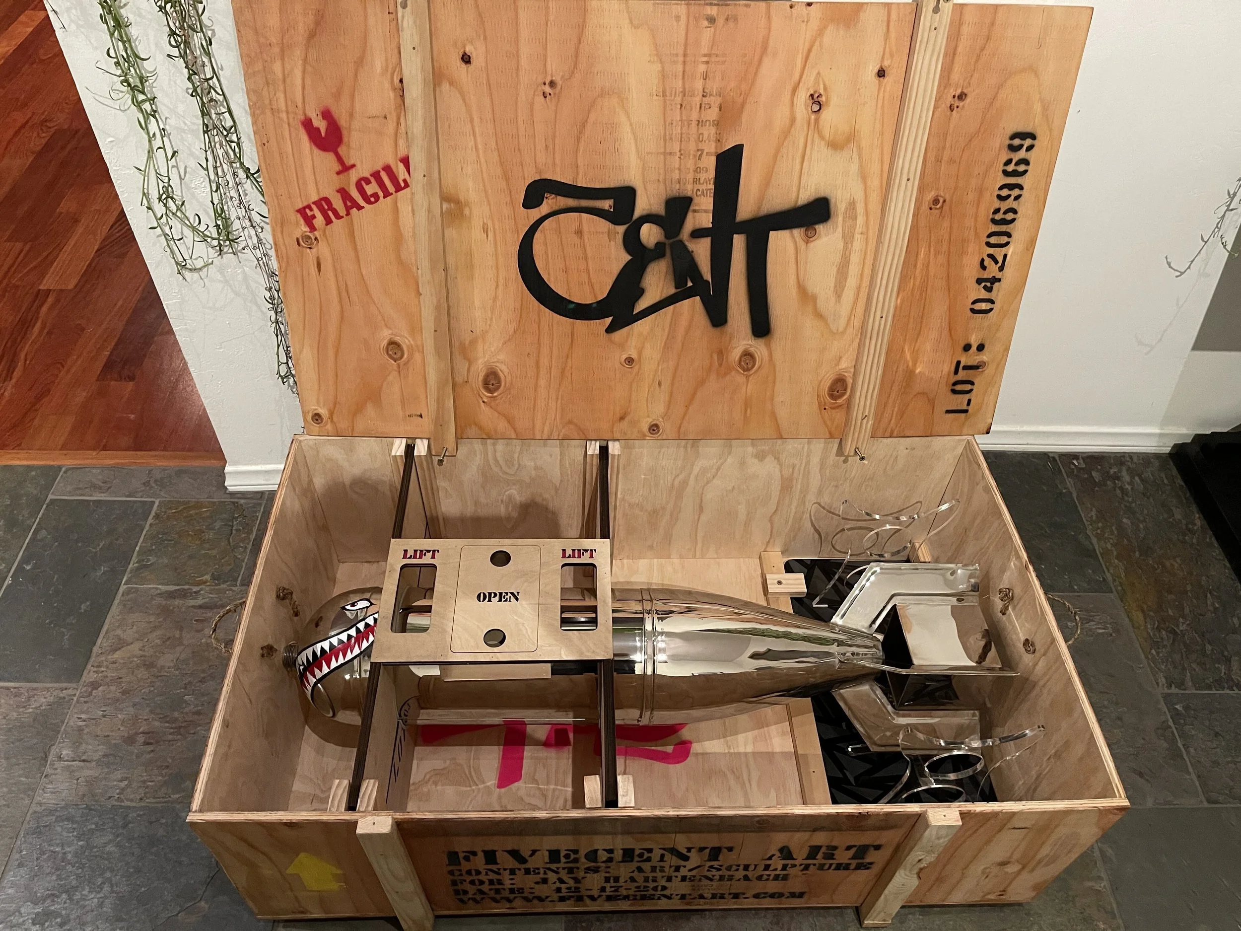 A large wooden crate containing a shiny metal rocket with a face painted on its nose, designed to look like a shark with sharp teeth. The crate has a lid propped open with the words "FUEL CONTRACT ART" and a black logo on the inside of the lid, along