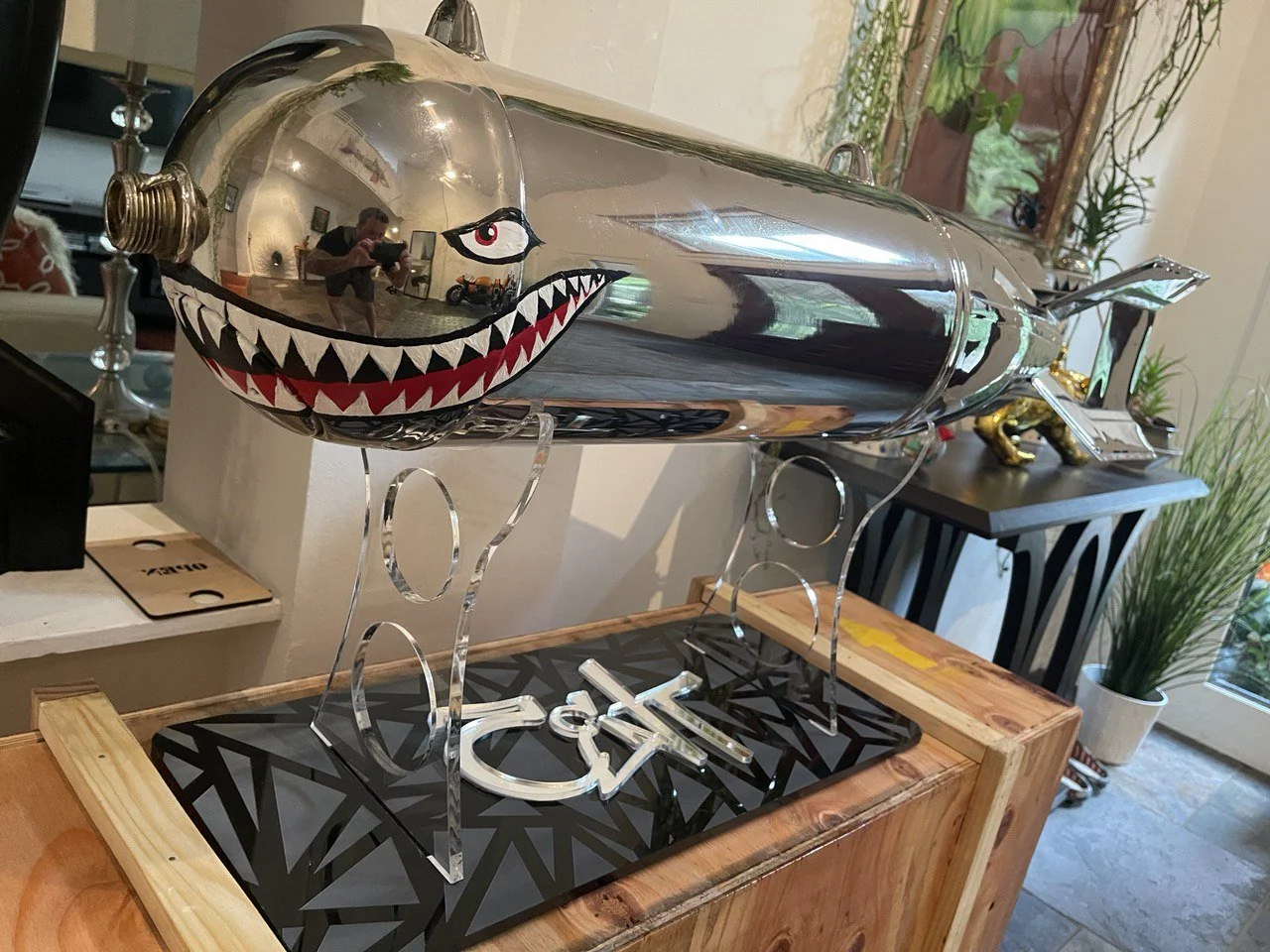 A decorative shark sculpture made from a metallic, reflective material, with painted eyes and sharp teeth, displayed on a black stand with an intricate geometric pattern, placed on top of a wooden surface.