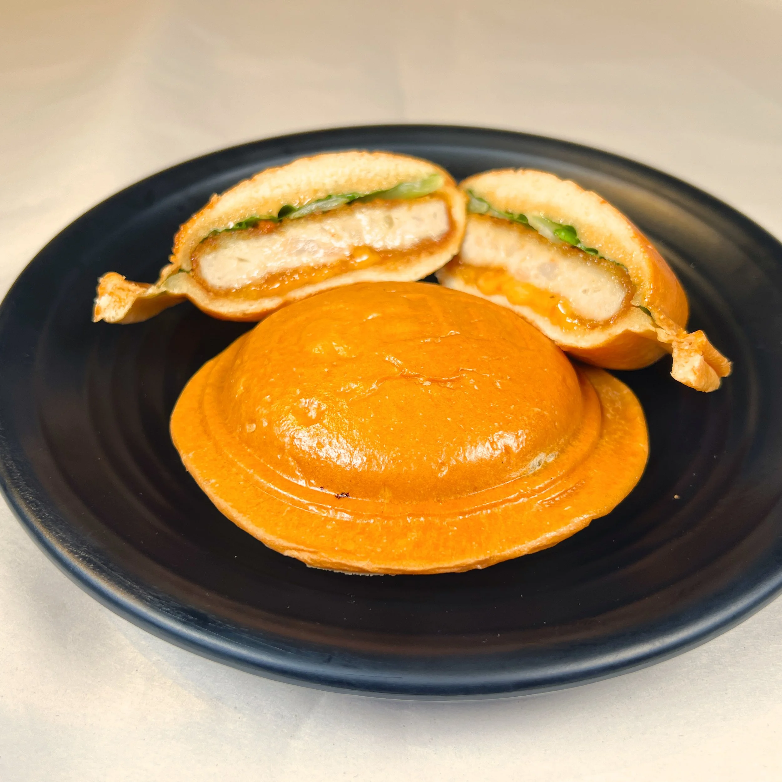 A sliced chicken sandwich on a black plate with lettuce and sauce, a whole sandwich bun in front.