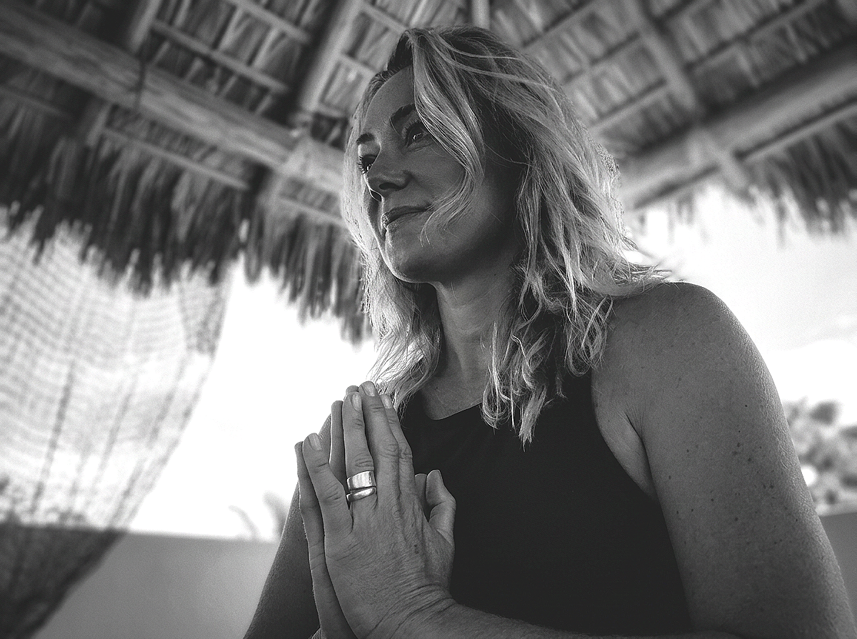 Woman practicing yoga outdoors under a thatched roof, hands in prayer position, with a serene expression.